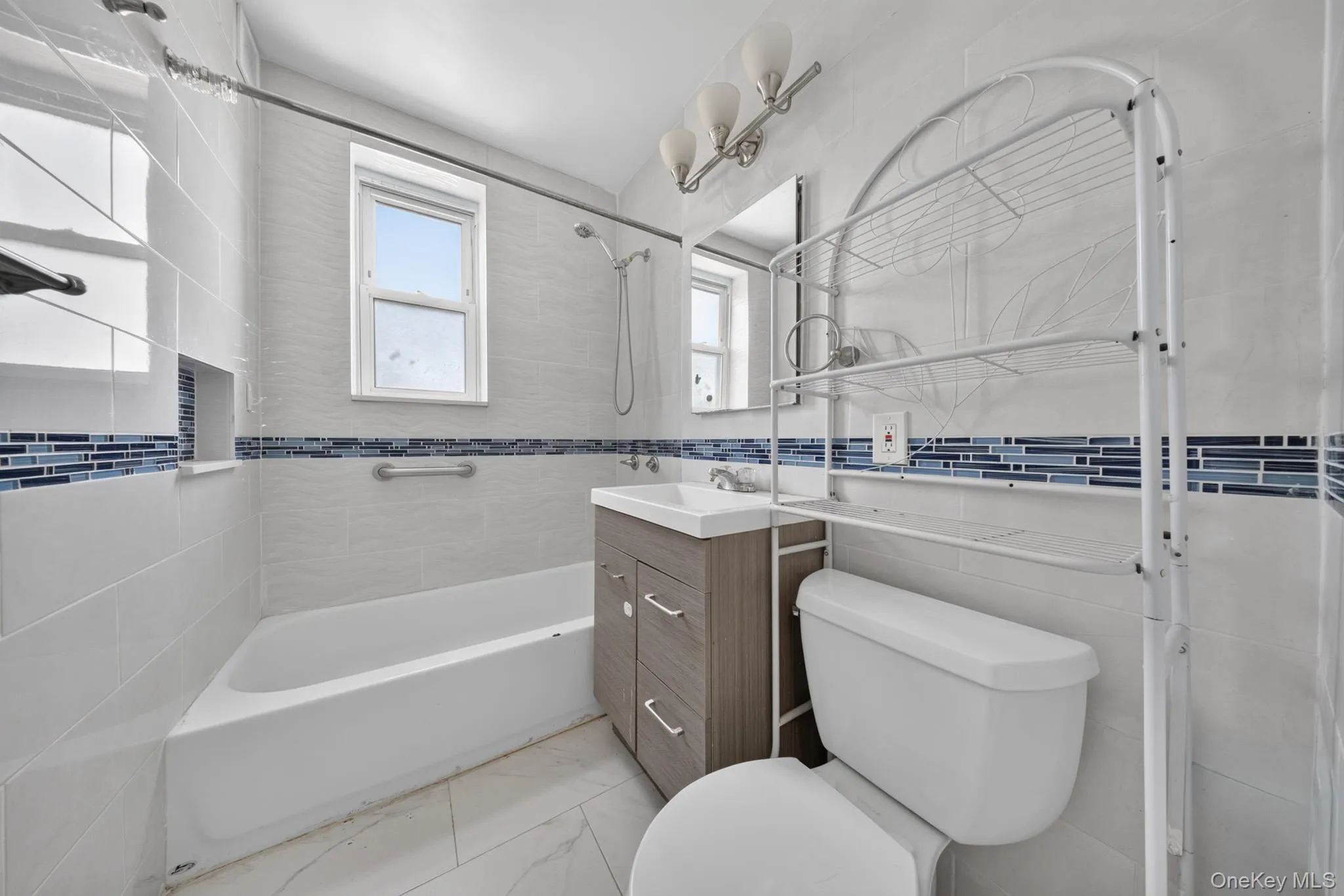 Bathroom featuring a white bathtub, tiled walls with a blue mosaic accent, a window, a vanity with under-sink storage, and a wall-mounted light fixture Bathroom featuring a white bathtub, tiled walls with a blue mosaic accent, a window, a vanity with under-sink storage, and a wall-mounted light fixture