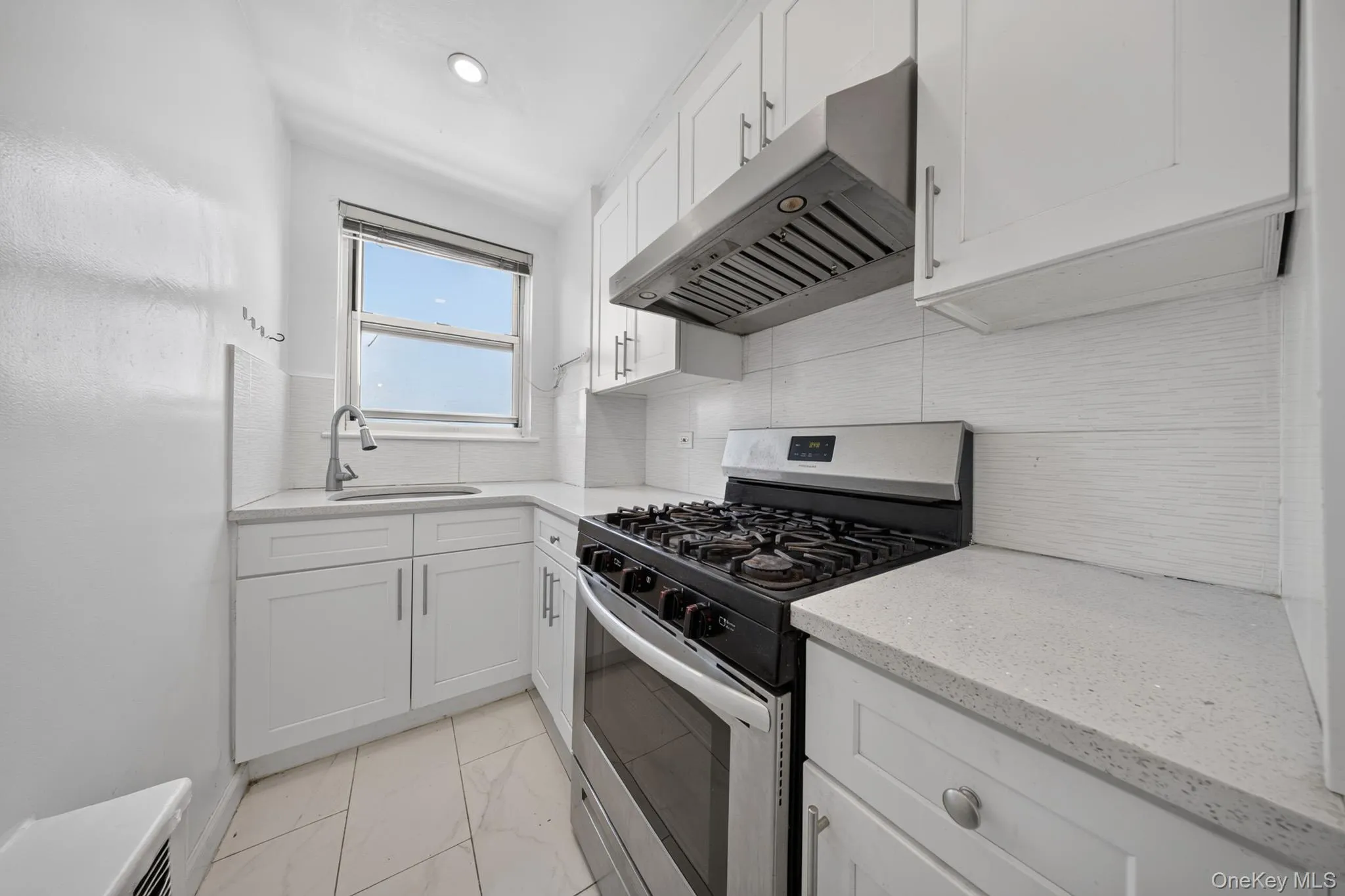 Modern kitchen featuring white cabinetry, light-toned countertops, a stainless steel gas range with coordinating hood, a stainless steel sink with a gooseneck faucet, and white tile flooring Modern kitchen featuring white cabinetry, light-toned countertops, a stainless steel gas range with coordinating hood, a stainless steel sink with a gooseneck faucet, and white tile flooring