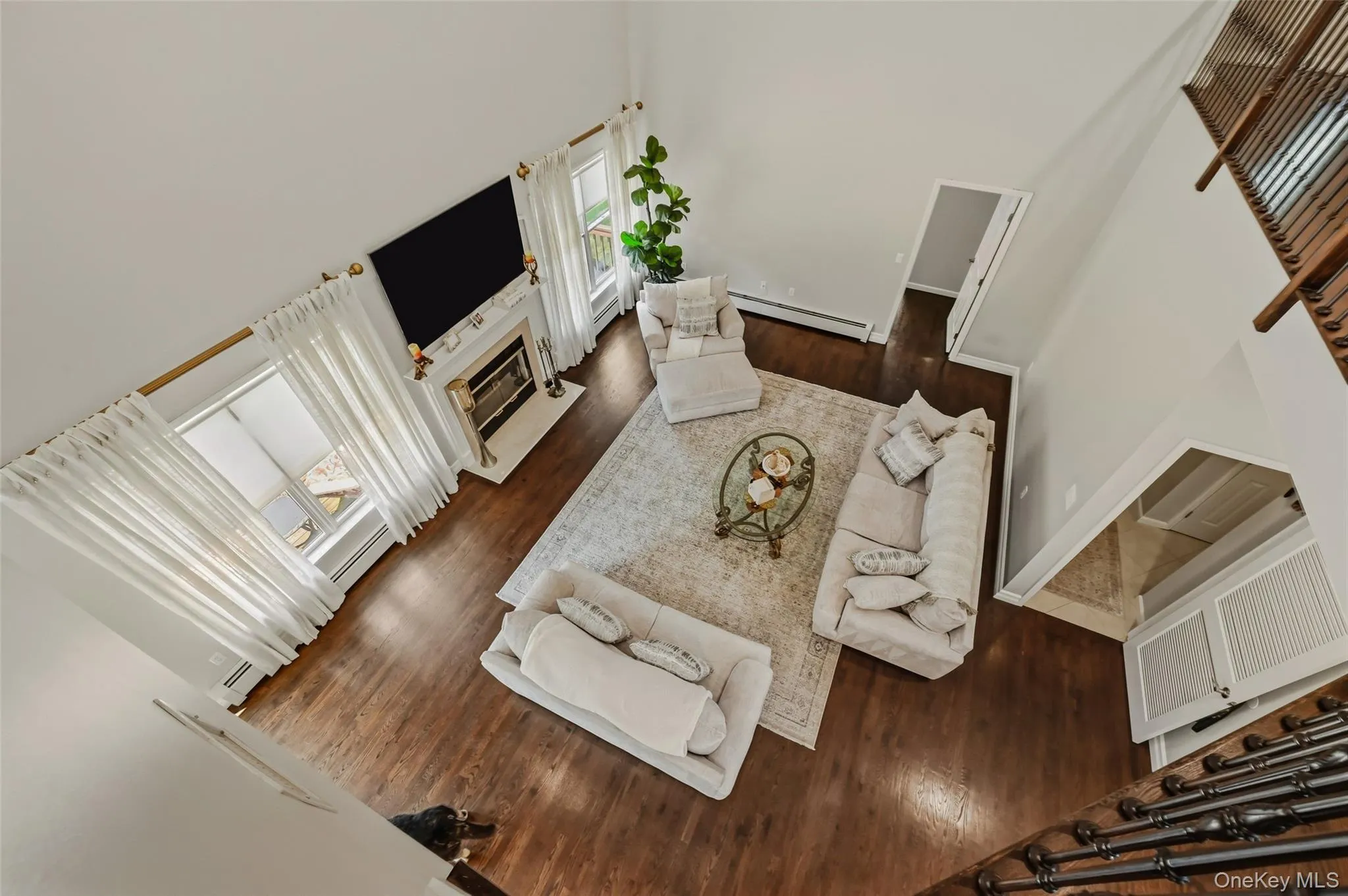 Two-story living area featuring wood-finish flooring, a white fireplace with a mantle, and large windows with white drapery Two-story living area featuring wood-finish flooring, a white fireplace with a mantle, and large windows with white drapery