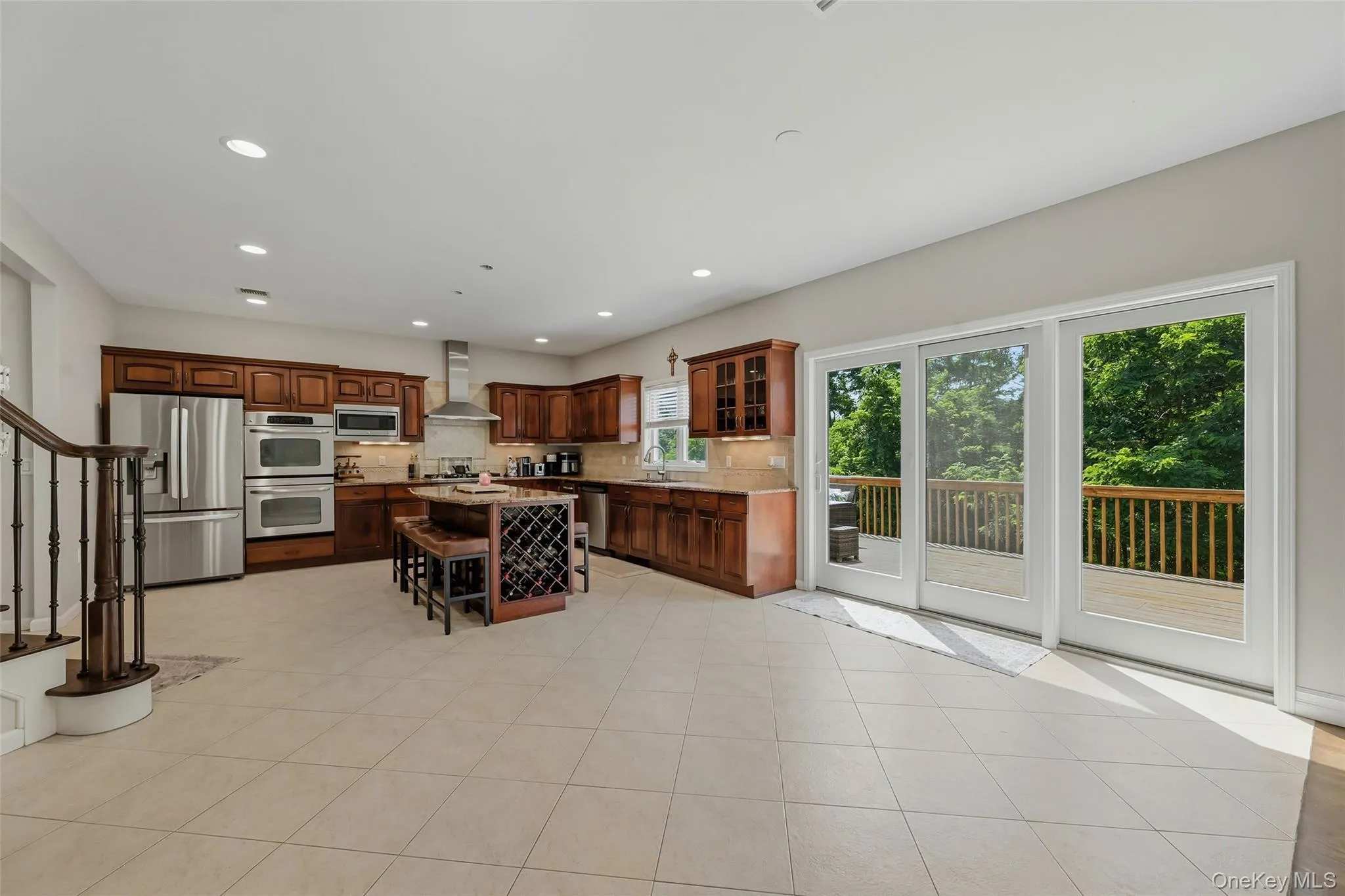 Expansive kitchen featuring wood cabinetry, stainless steel appliances including double ovens, a central island with integrated wine storage, and direct access to an outdoor deck through glass sliding doors Expansive kitchen featuring wood cabinetry, stainless steel appliances including double ovens, a central island with integrated wine storage, and direct access to an outdoor deck through glass sliding doors