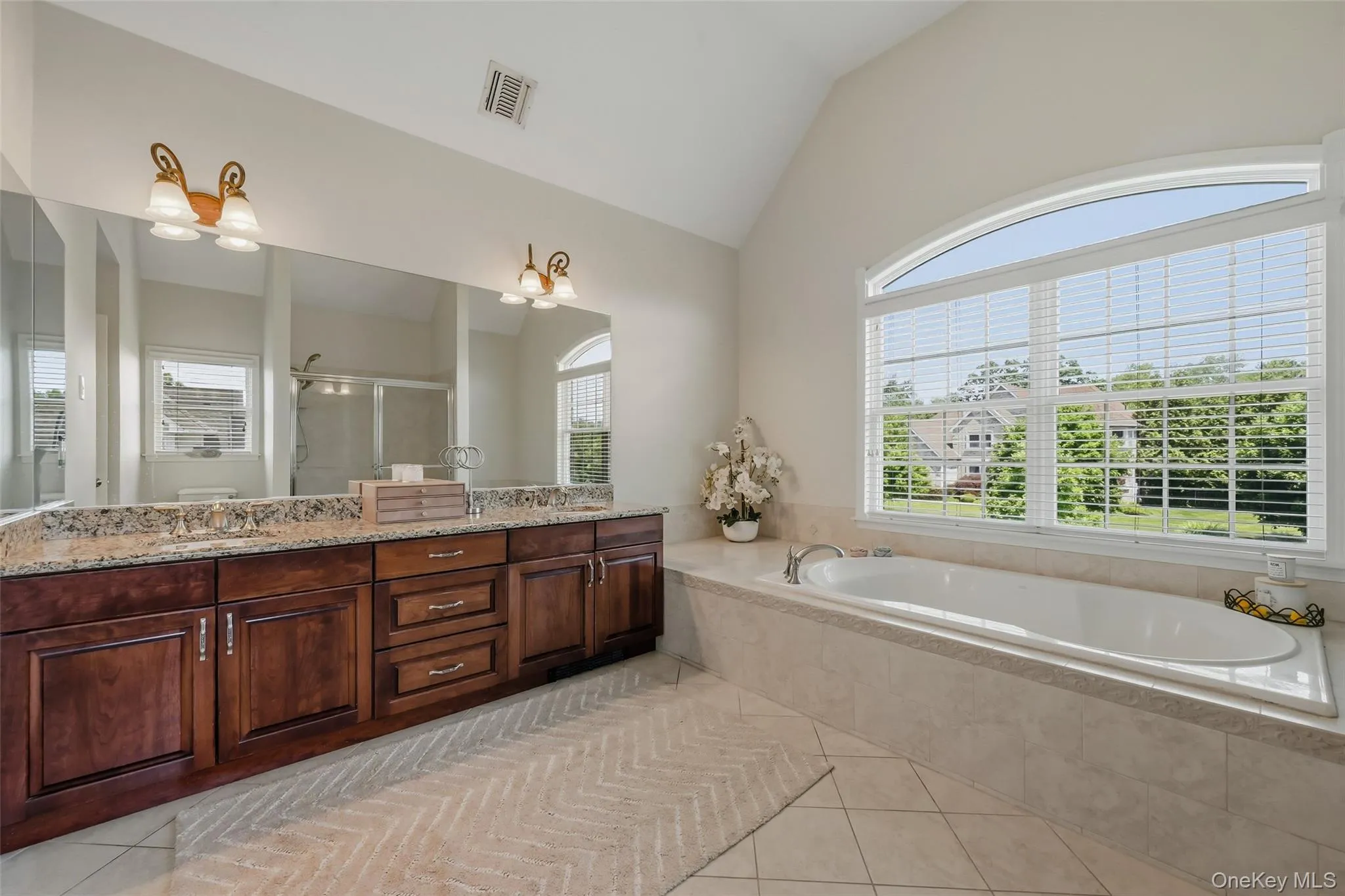 Spacious bathroom featuring a large soaking tub with tiled surround, a double vanity with stone countertop, and an expansive arched window Spacious bathroom featuring a large soaking tub with tiled surround, a double vanity with stone countertop, and an expansive arched window
