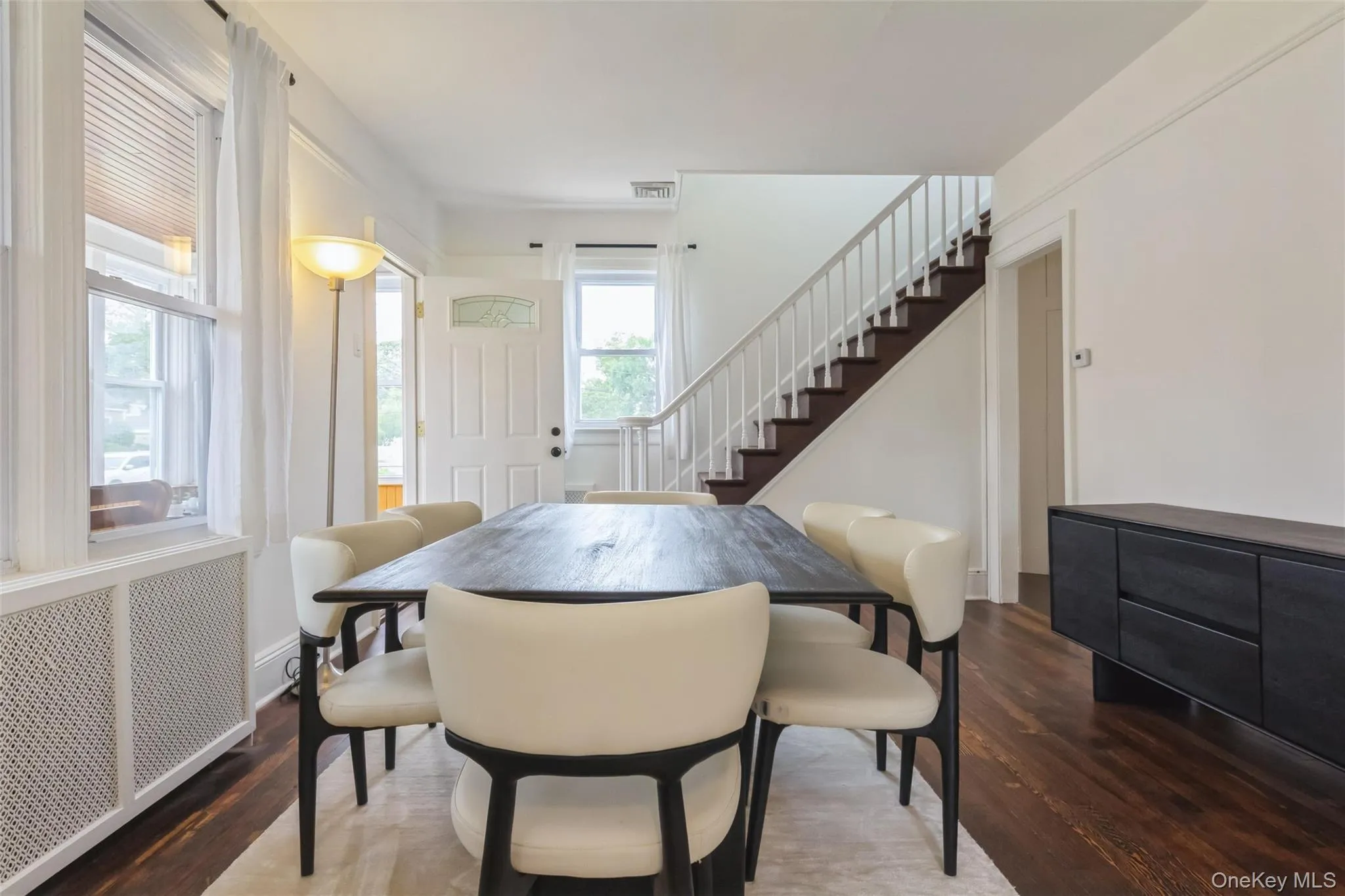 Dining area with rich wood-finish flooring and a prominent staircase featuring white risers and railings Dining area with rich wood-finish flooring and a prominent staircase featuring white risers and railings