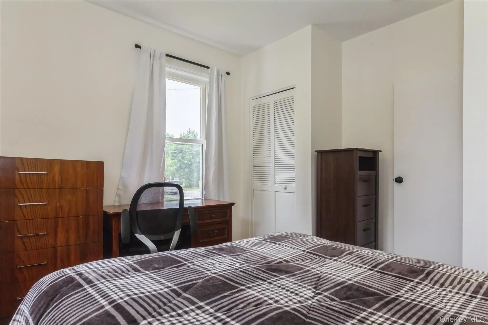 Bright room featuring a window with white curtains, white walls, and a white bi-fold closet door Bright room featuring a window with white curtains, white walls, and a white bi-fold closet door