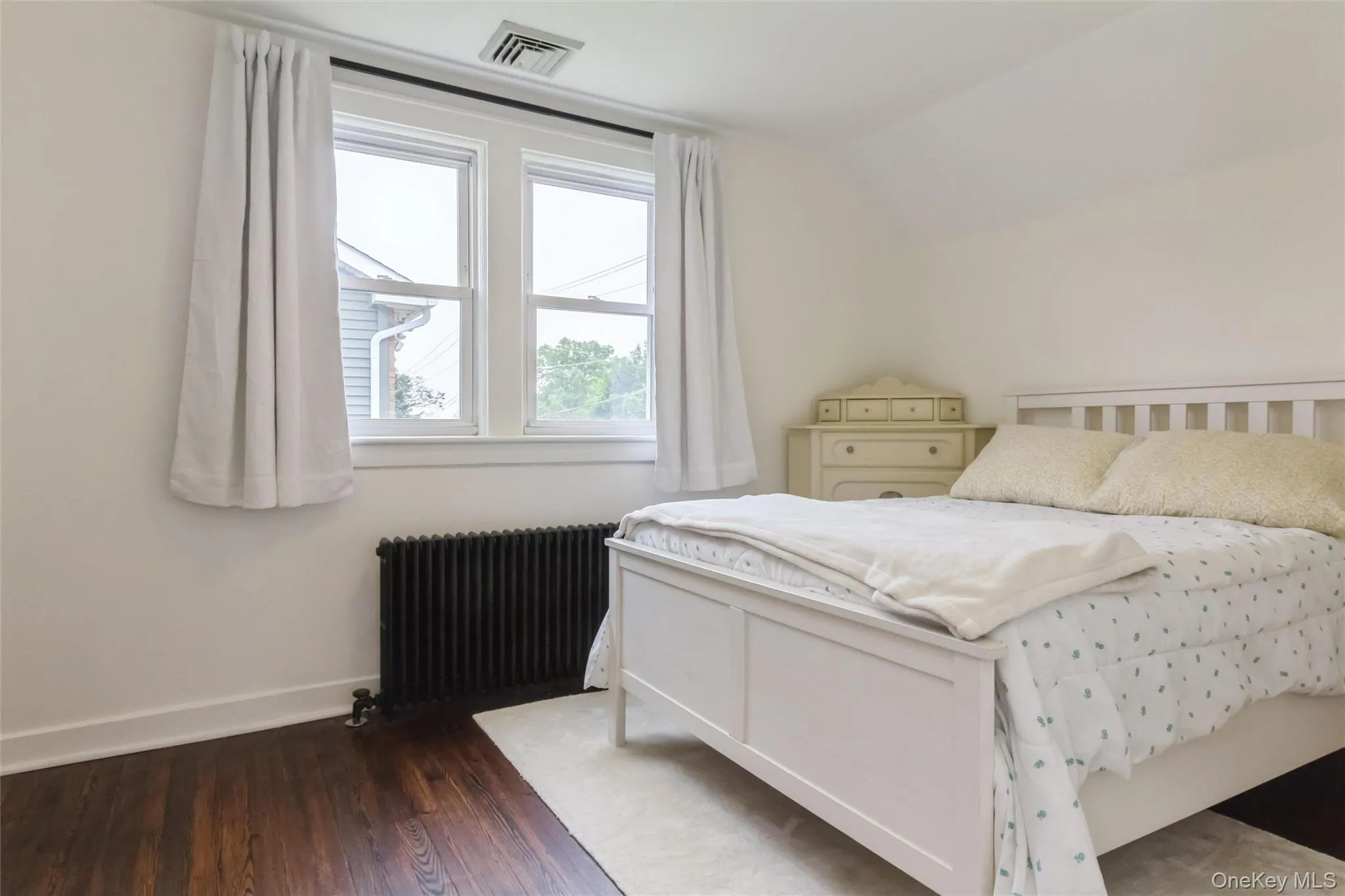 Bedroom featuring dark wood-finish flooring, white walls, and a prominent window with white trim Bedroom featuring dark wood-finish flooring, white walls, and a prominent window with white trim