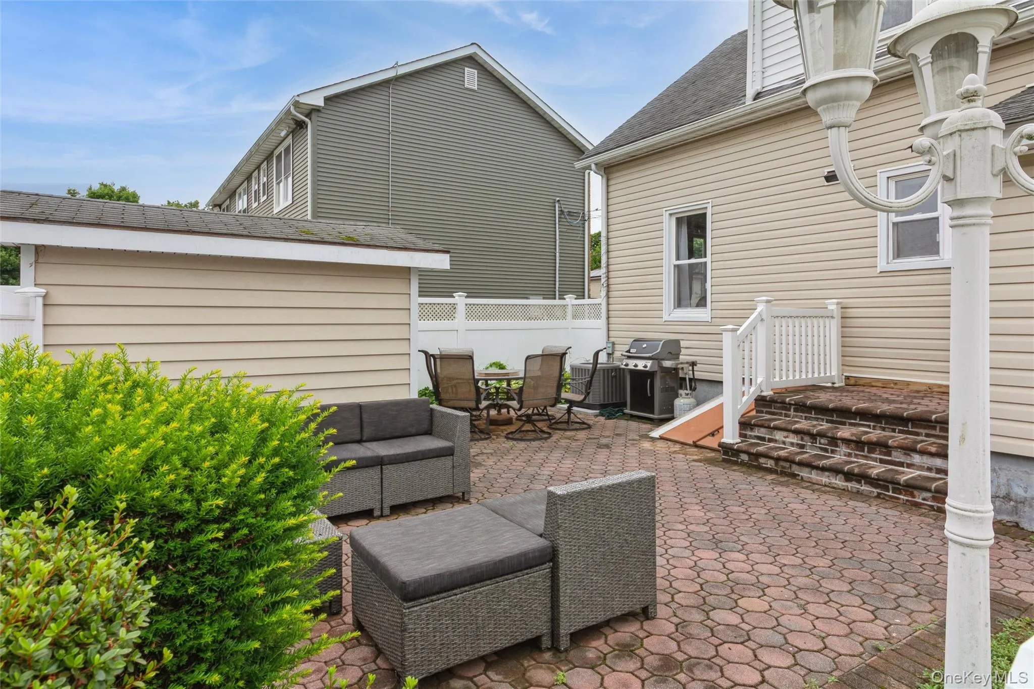 Brick-patterned paver patio featuring a decorative white privacy fence, multi-level brick steps with white railings, and mature shrubbery Brick-patterned paver patio featuring a decorative white privacy fence, multi-level brick steps with white railings, and mature shrubbery