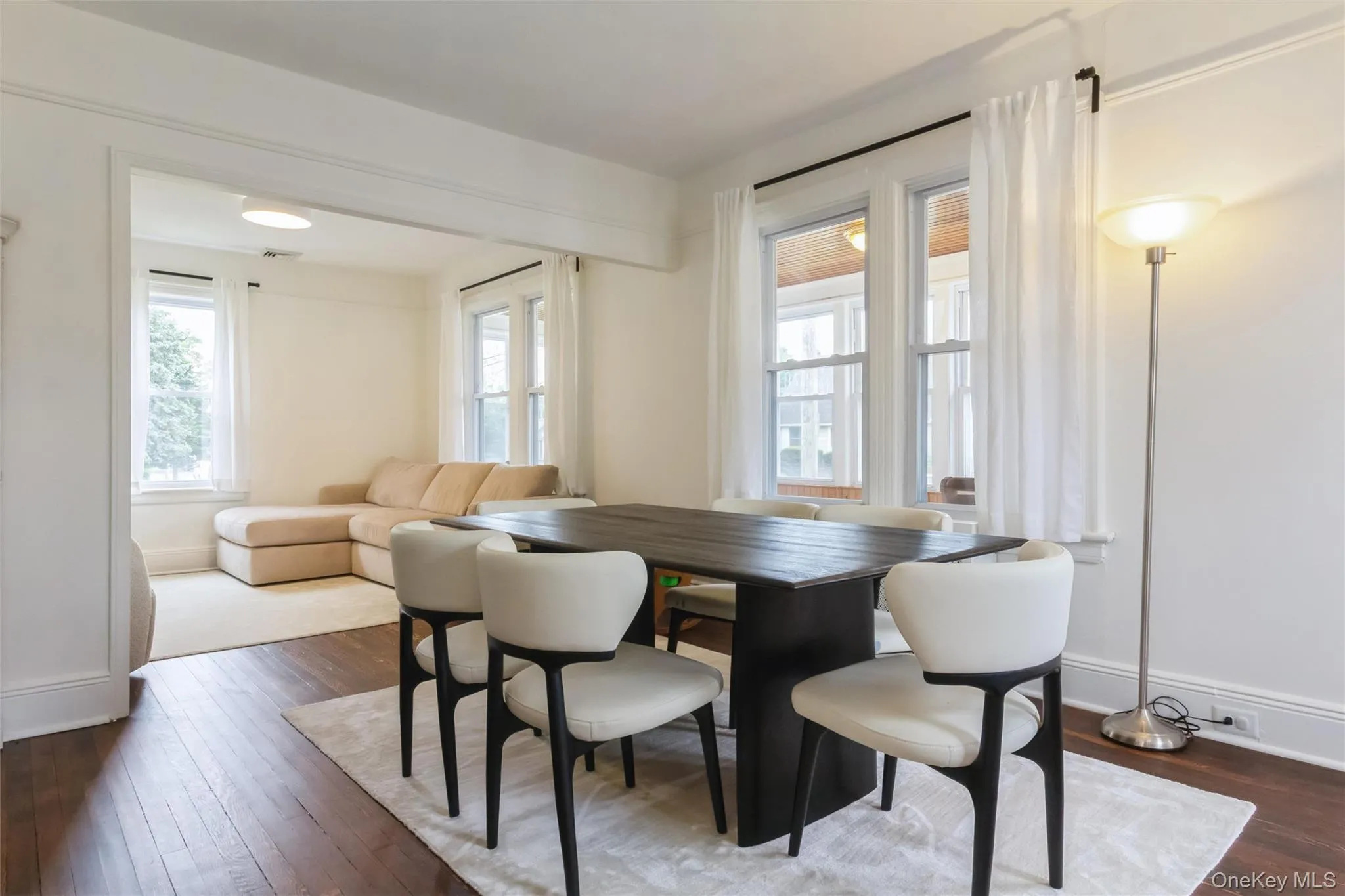 Open concept living space featuring rich wood-finish flooring, white trim, and multiple windows with sheer white drapery Open concept living space featuring rich wood-finish flooring, white trim, and multiple windows with sheer white drapery