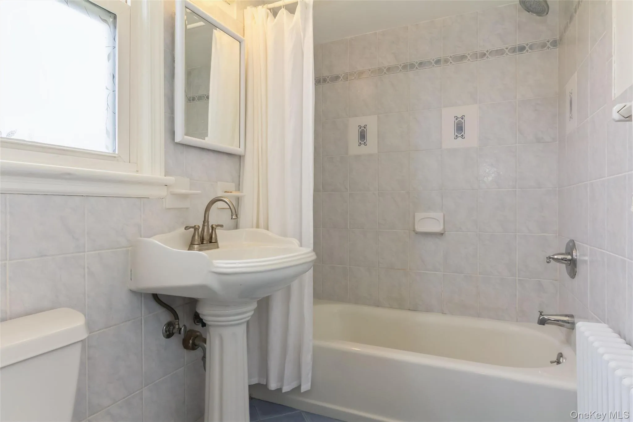 Bathroom featuring a pedestal sink with brushed nickel faucet, a white bathtub with tile surround, a built-in shower head, and a window with privacy glass Bathroom featuring a pedestal sink with brushed nickel faucet, a white bathtub with tile surround, a built-in shower head, and a window with privacy glass