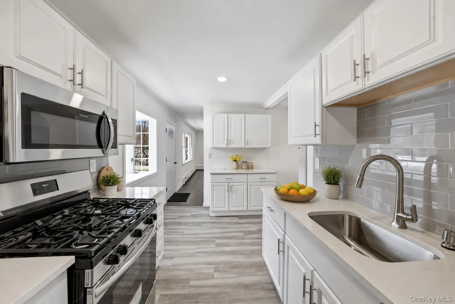 Modern kitchen featuring white shaker cabinetry, stainless steel appliances including a gas range and microwave, light gray subway tile backsplash, and wood-finish flooring Modern kitchen featuring white shaker cabinetry, stainless steel appliances including a gas range and microwave, light gray subway tile backsplash, and wood-finish flooring