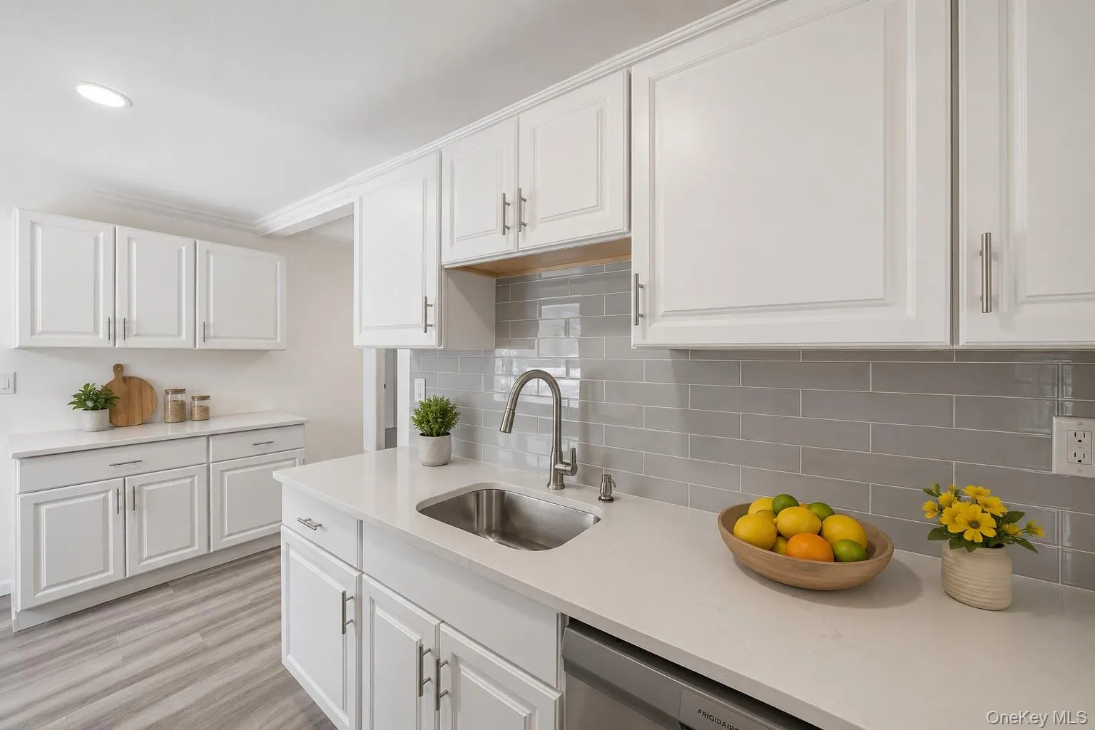 Bright kitchen featuring white shaker cabinetry, light-toned countertops, a subway tile backsplash, and wood-finish flooring Bright kitchen featuring white shaker cabinetry, light-toned countertops, a subway tile backsplash, and wood-finish flooring