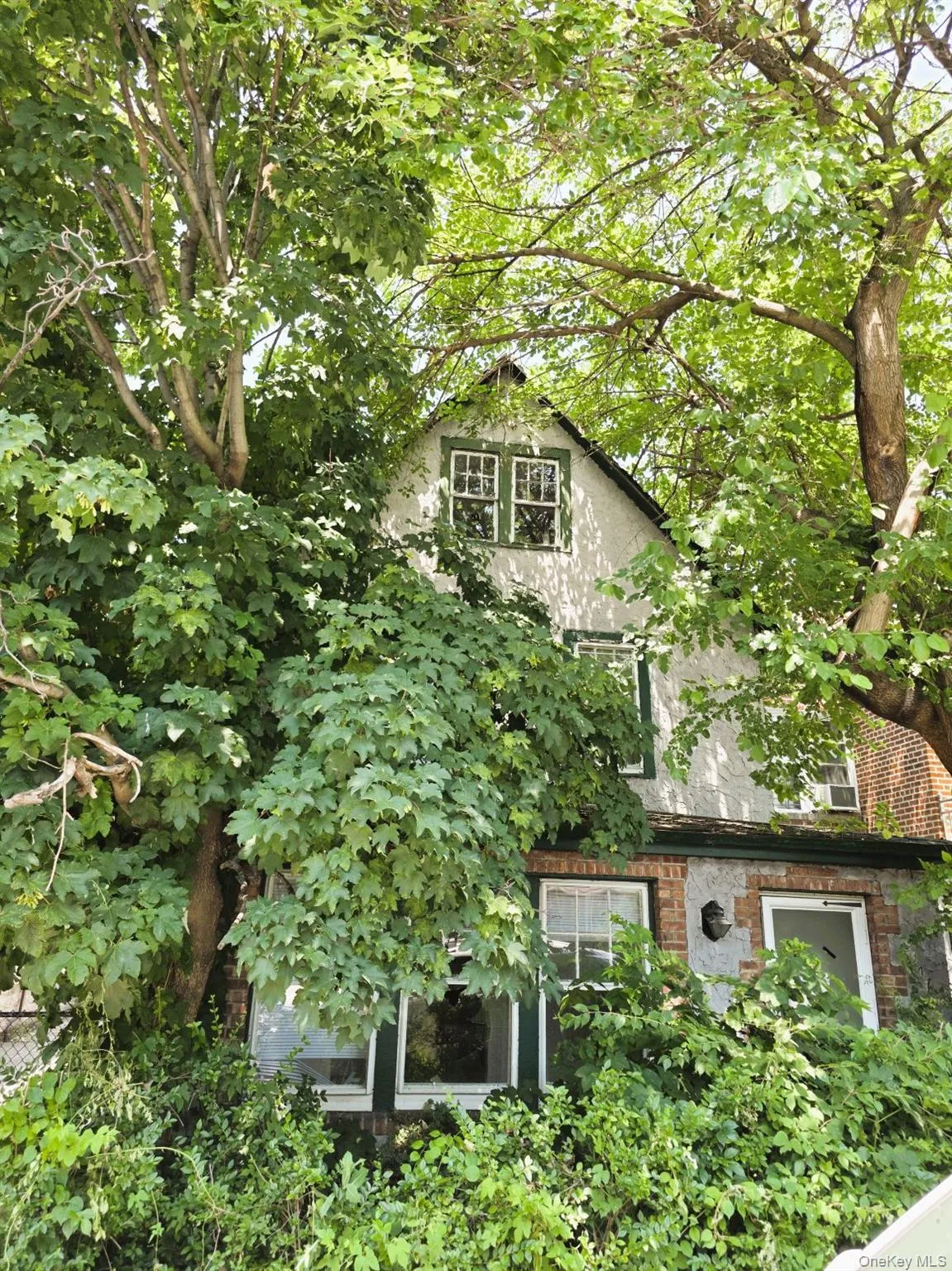 Exterior featuring mature trees, a stucco facade, brick accents, and white-framed windows with dark green trim Exterior featuring mature trees, a stucco facade, brick accents, and white-framed windows with dark green trim