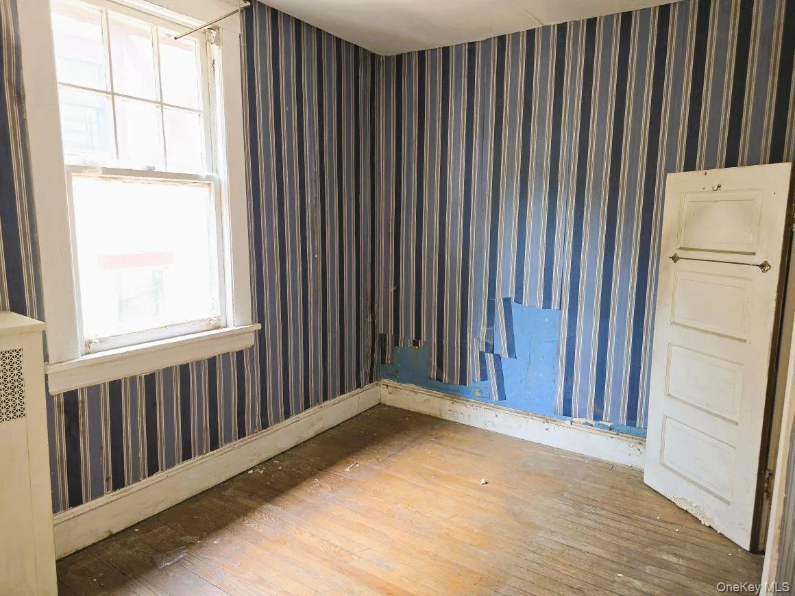 Room featuring wood-finish flooring, striped wallpaper, a double-hung window with white trim, a white baseboard, and a white interior door Room featuring wood-finish flooring, striped wallpaper, a double-hung window with white trim, a white baseboard, and a white interior door
