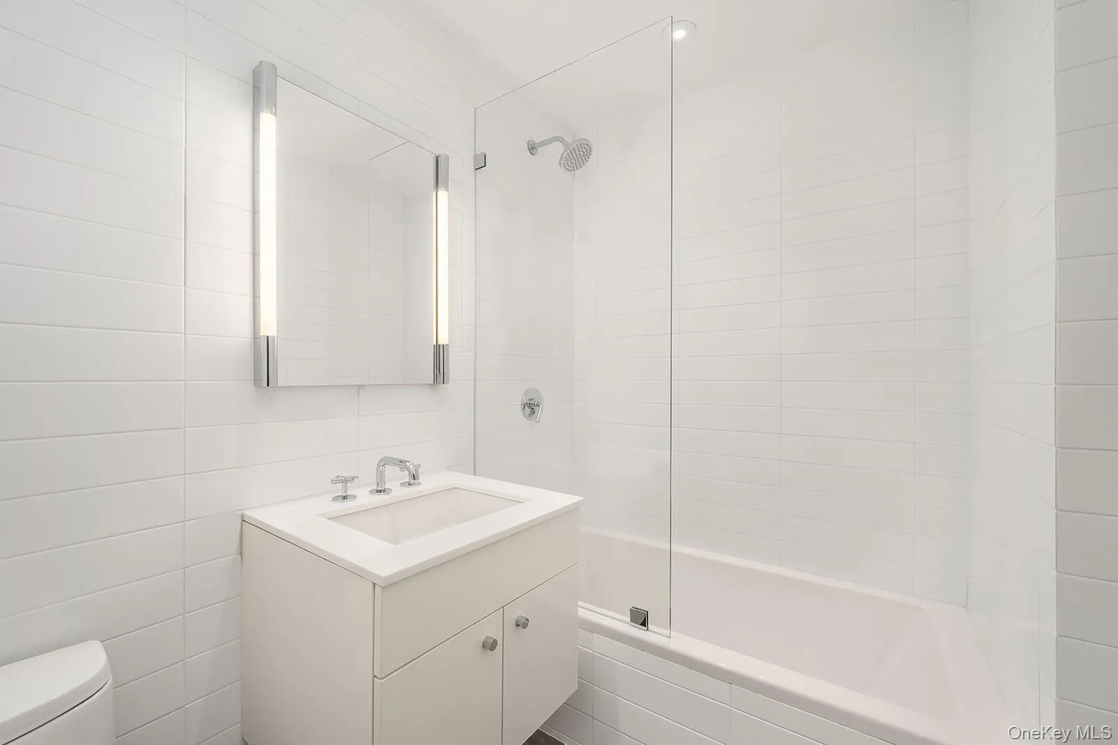 Bathroom featuring a vanity with an integrated sink and chrome fixtures, illuminated mirror with vertical lighting, floor-to-ceiling white tiled walls, and a bathtub with a clear glass shower screen and a chrome shower head Bathroom featuring a vanity with an integrated sink and chrome fixtures, illuminated mirror with vertical lighting, floor-to-ceiling white tiled walls, and a bathtub with a clear glass shower screen and a chrome shower head