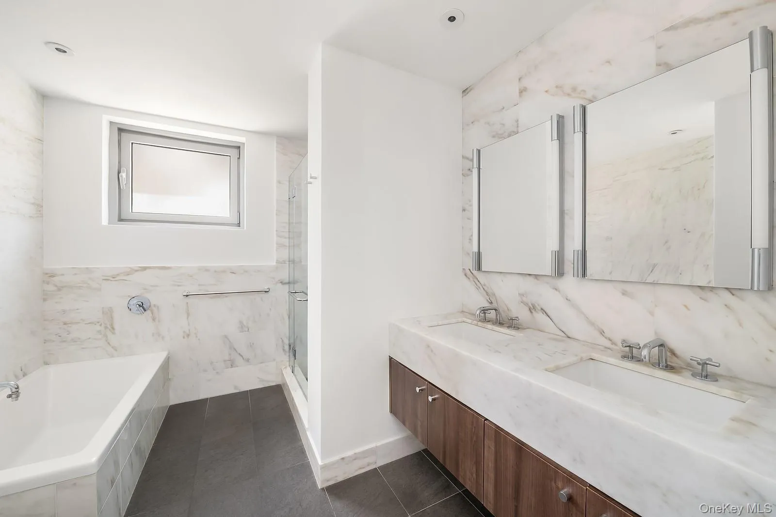 Bathroom featuring a floating double vanity with integrated sinks and a wood-finish base, dual wall-mounted illuminated mirrors, and a full-height tile backsplash Bathroom featuring a floating double vanity with integrated sinks and a wood-finish base, dual wall-mounted illuminated mirrors, and a full-height tile backsplash