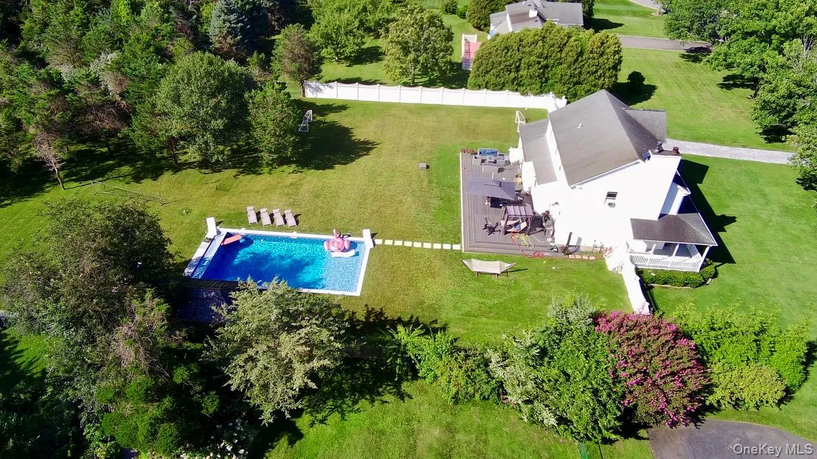 Expansive green lawn featuring an in-ground swimming pool, a large outdoor patio, mature trees, and a white picket fence. Expansive green lawn featuring an in-ground swimming pool, a large outdoor patio, mature trees, and a white picket fence.