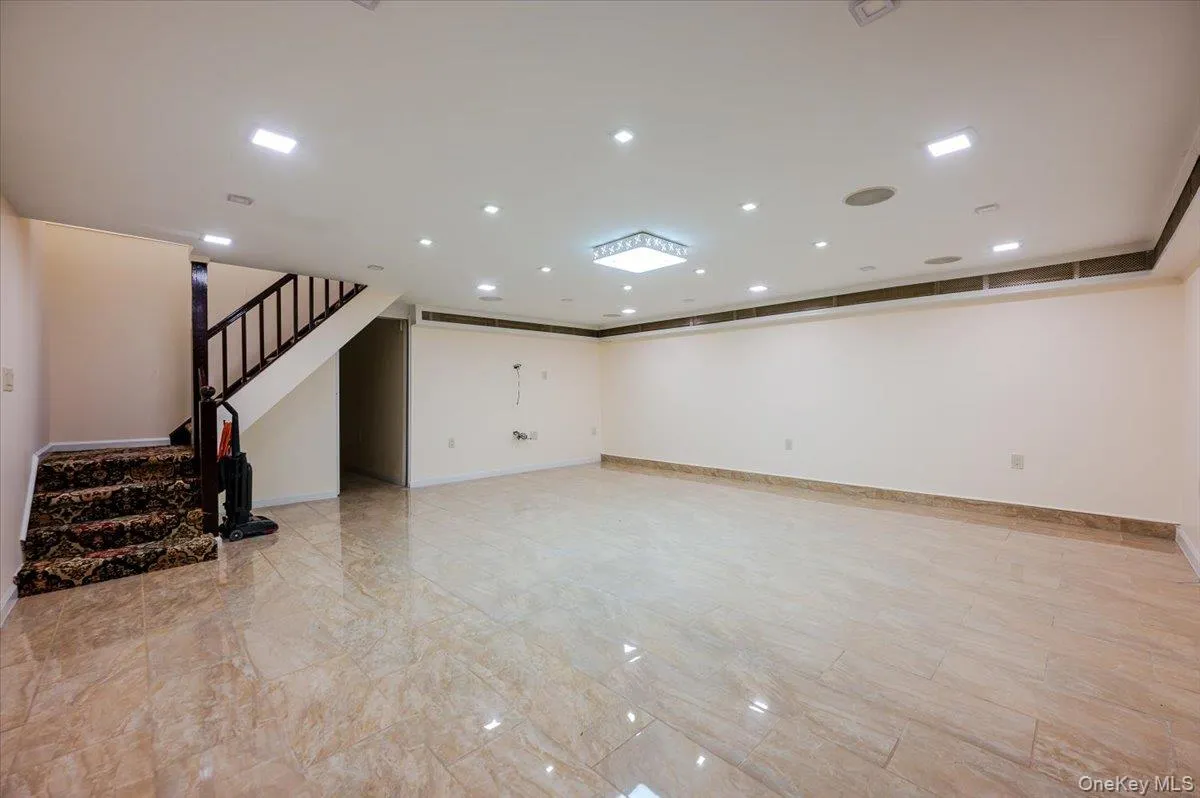 Spacious room featuring polished tile flooring, recessed ceiling lighting, a contemporary square ceiling fixture, and a staircase with a dark wood-finish railing Spacious room featuring polished tile flooring, recessed ceiling lighting, a contemporary square ceiling fixture, and a staircase with a dark wood-finish railing
