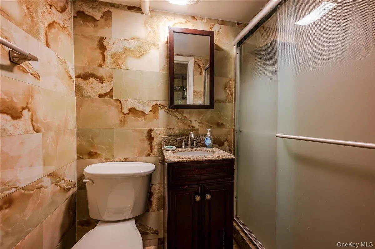 Bathroom featuring full-height tiled walls, a dark wood-finish vanity with a granite-style countertop, a white porcelain sink with a chrome faucet, and a frameless glass shower enclosure with a frosted panel Bathroom featuring full-height tiled walls, a dark wood-finish vanity with a granite-style countertop, a white porcelain sink with a chrome faucet, and a frameless glass shower enclosure with a frosted panel