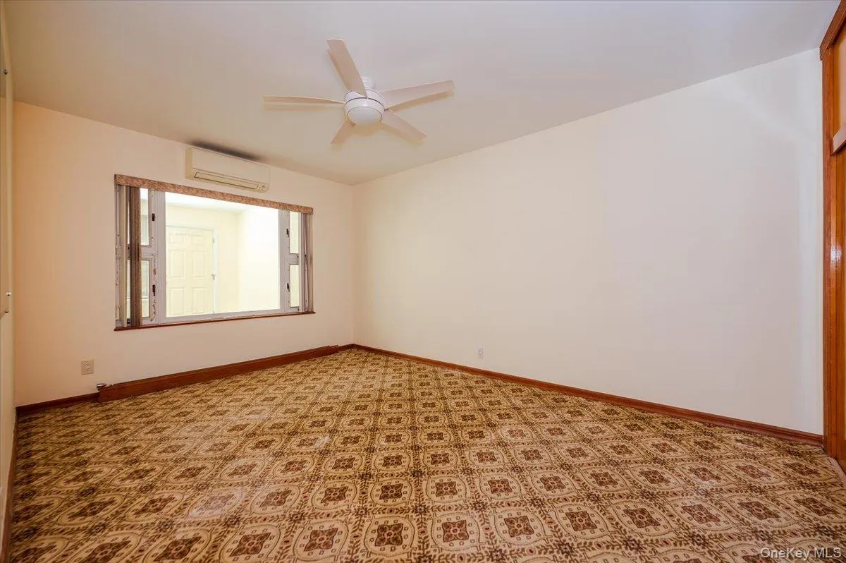 Spacious room featuring patterned carpeting, white walls, and a window with wooden framing Spacious room featuring patterned carpeting, white walls, and a window with wooden framing