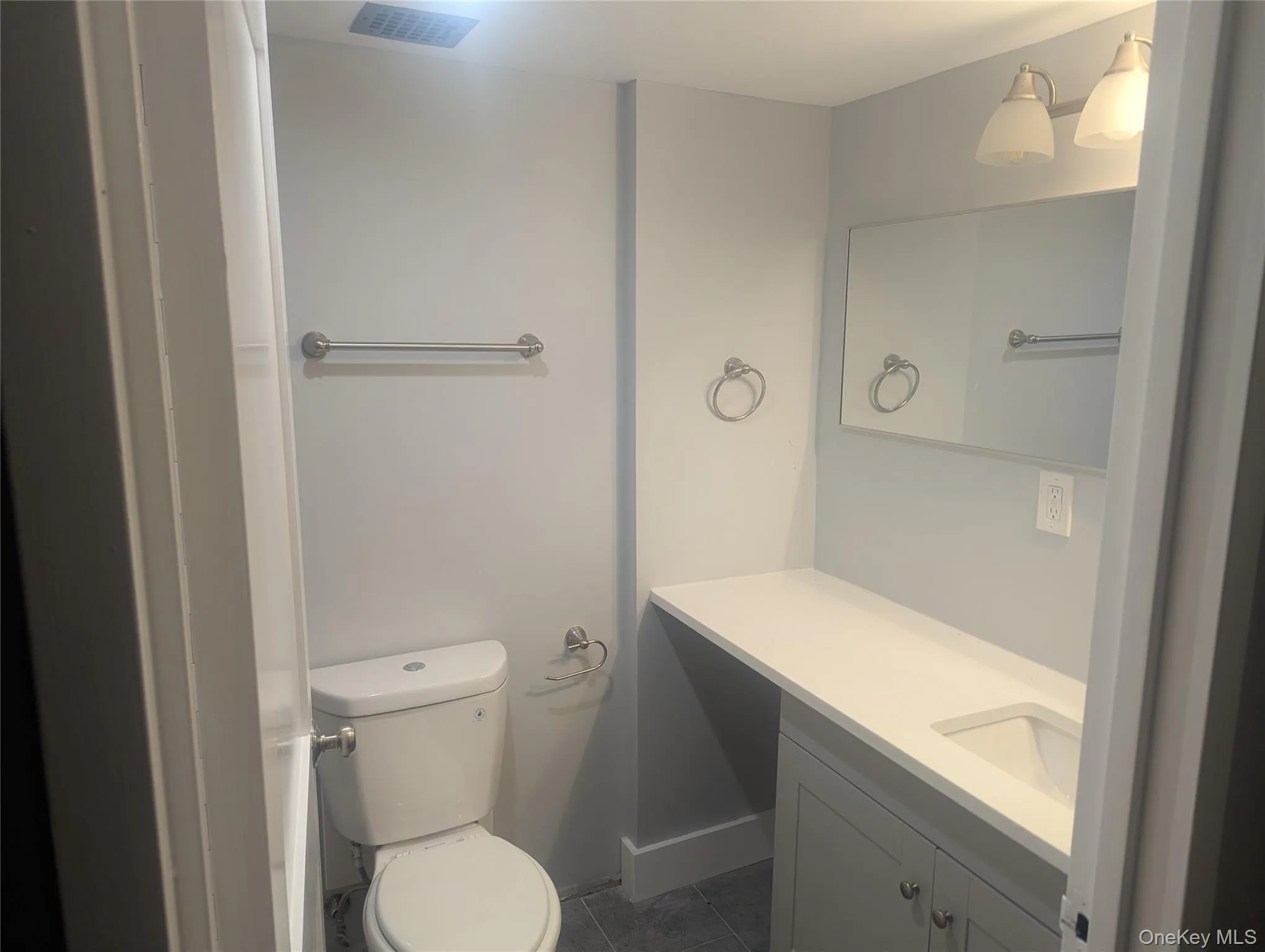 Bathroom featuring a white toilet, gray vanity with a built-in sink, white countertop, and wall-mounted mirror Bathroom featuring a white toilet, gray vanity with a built-in sink, white countertop, and wall-mounted mirror