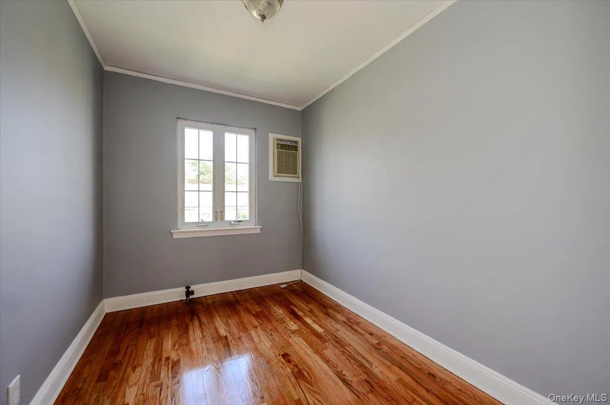 Hardwood flooring, light gray wall finish, window with white trim, white ceiling with crown molding, and a window-mounted air conditioning unit Hardwood flooring, light gray wall finish, window with white trim, white ceiling with crown molding, and a window-mounted air conditioning unit