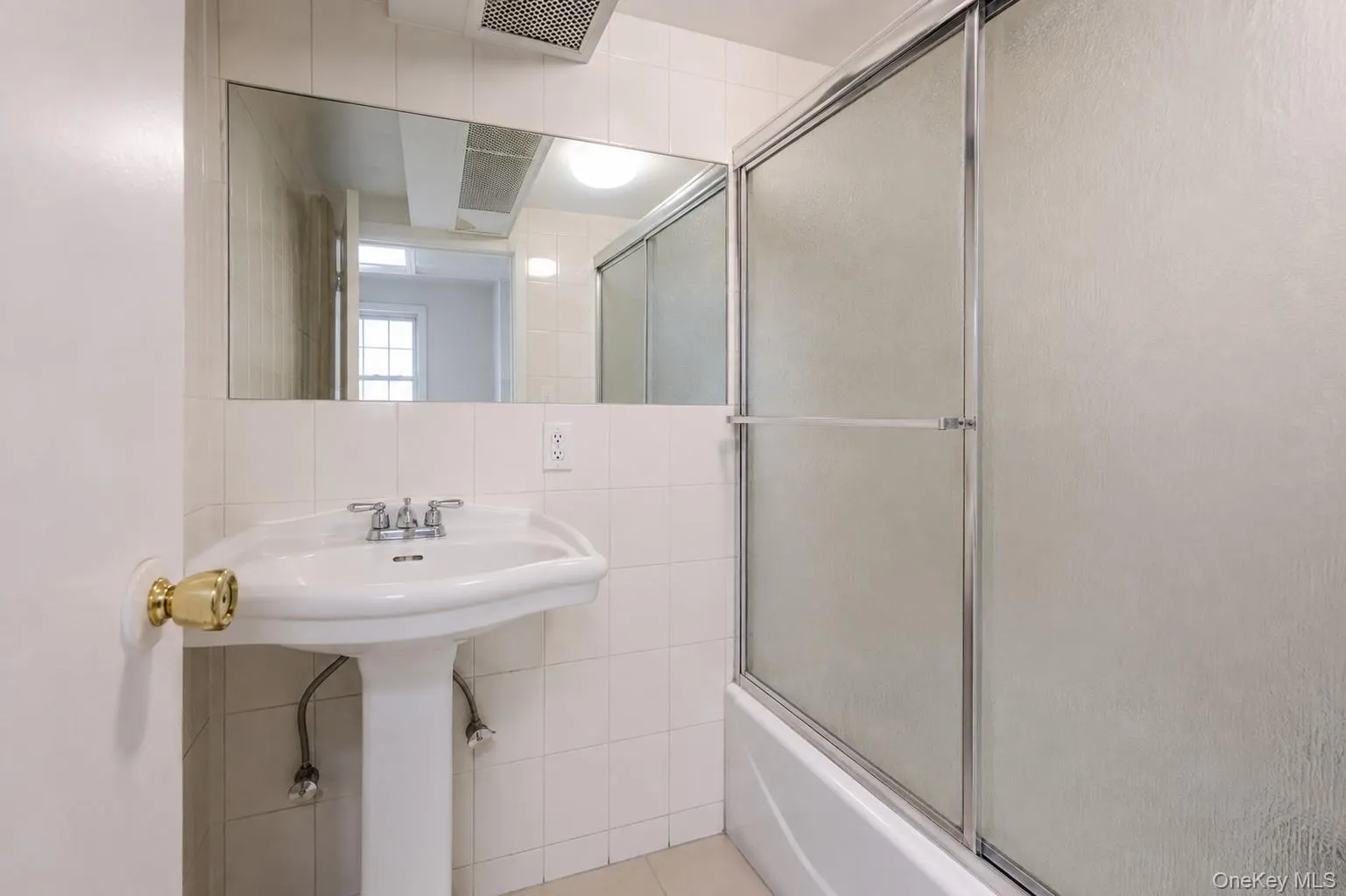 White tile walls, pedestal sink, chrome faucet, wall-mounted mirror, and a bathtub with sliding glass doors White tile walls, pedestal sink, chrome faucet, wall-mounted mirror, and a bathtub with sliding glass doors