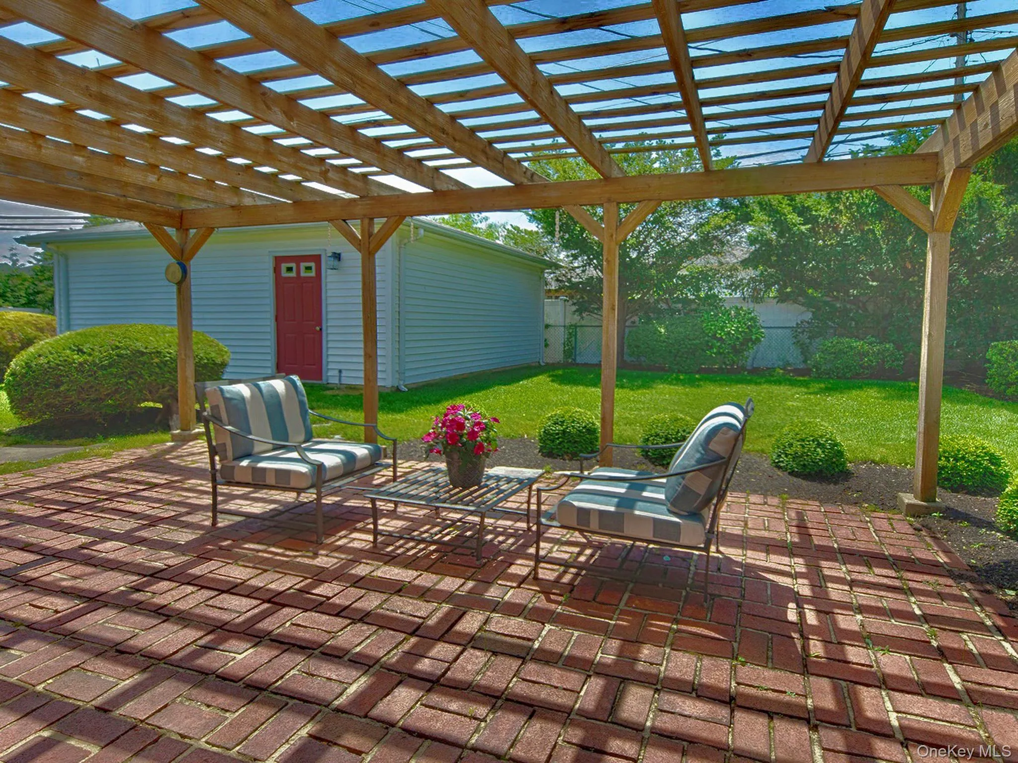 Brick paver patio featuring a wooden pergola with translucent roofing panels, surrounded by a manicured lawn, mature shrubs, and fencing Brick paver patio featuring a wooden pergola with translucent roofing panels, surrounded by a manicured lawn, mature shrubs, and fencing