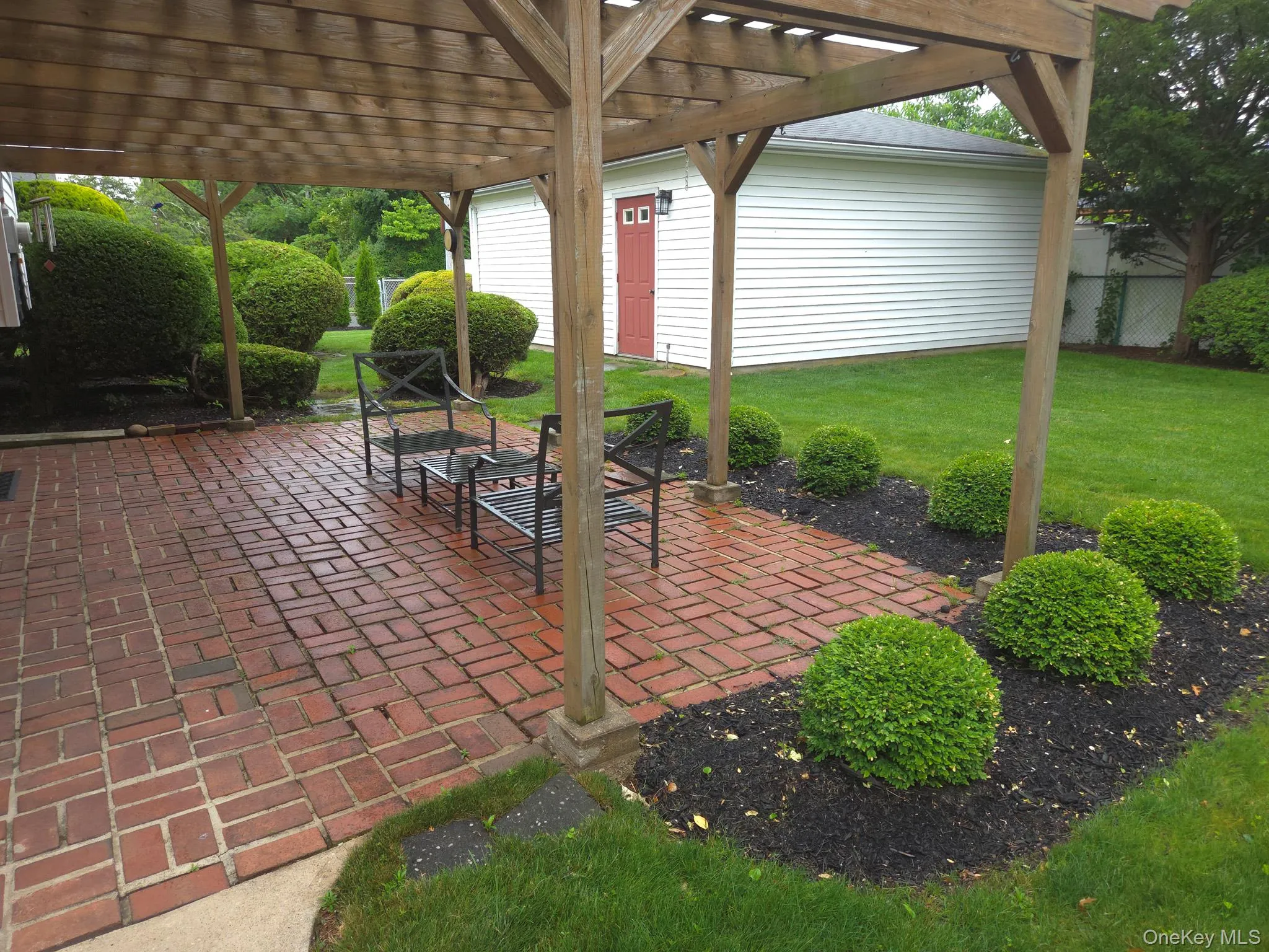 Brick paver patio featuring a wooden pergola with translucent roofing panels, surrounded by a manicured lawn, mature shrubs, and fencing Brick paver patio featuring a wooden pergola with translucent roofing panels, surrounded by a manicured lawn, mature shrubs, and fencing