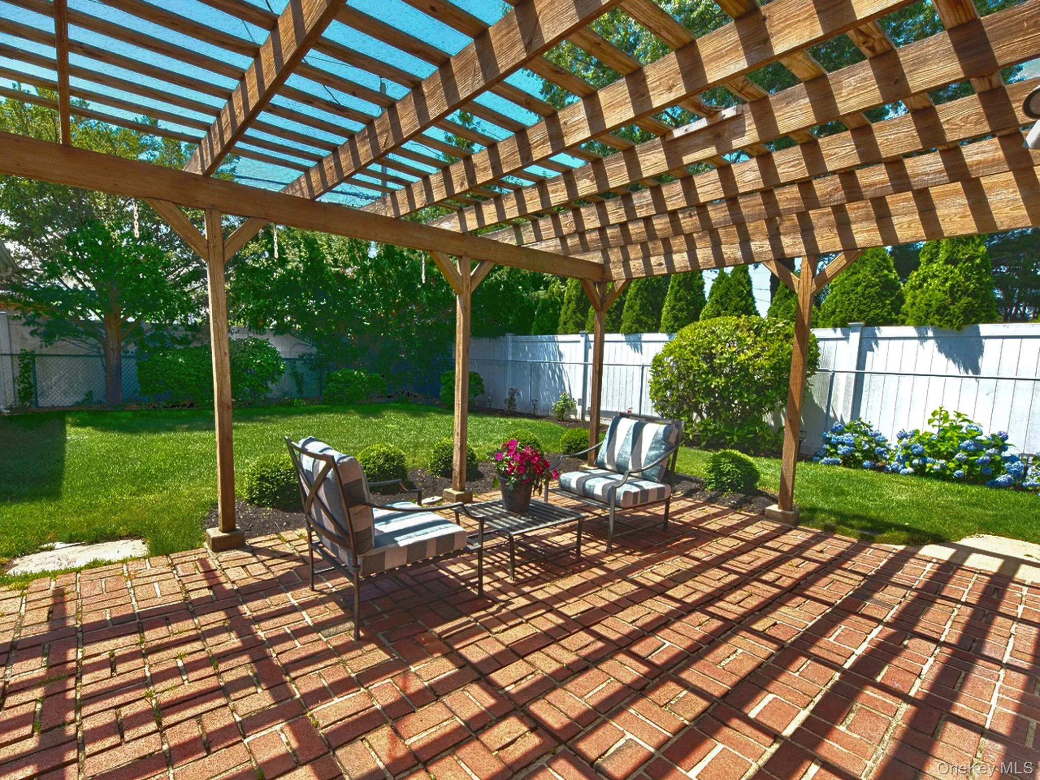 Brick paver patio featuring a wooden pergola with translucent roofing panels, surrounded by a manicured lawn, mature shrubs, and fencing Brick paver patio featuring a wooden pergola with translucent roofing panels, surrounded by a manicured lawn, mature shrubs, and fencing