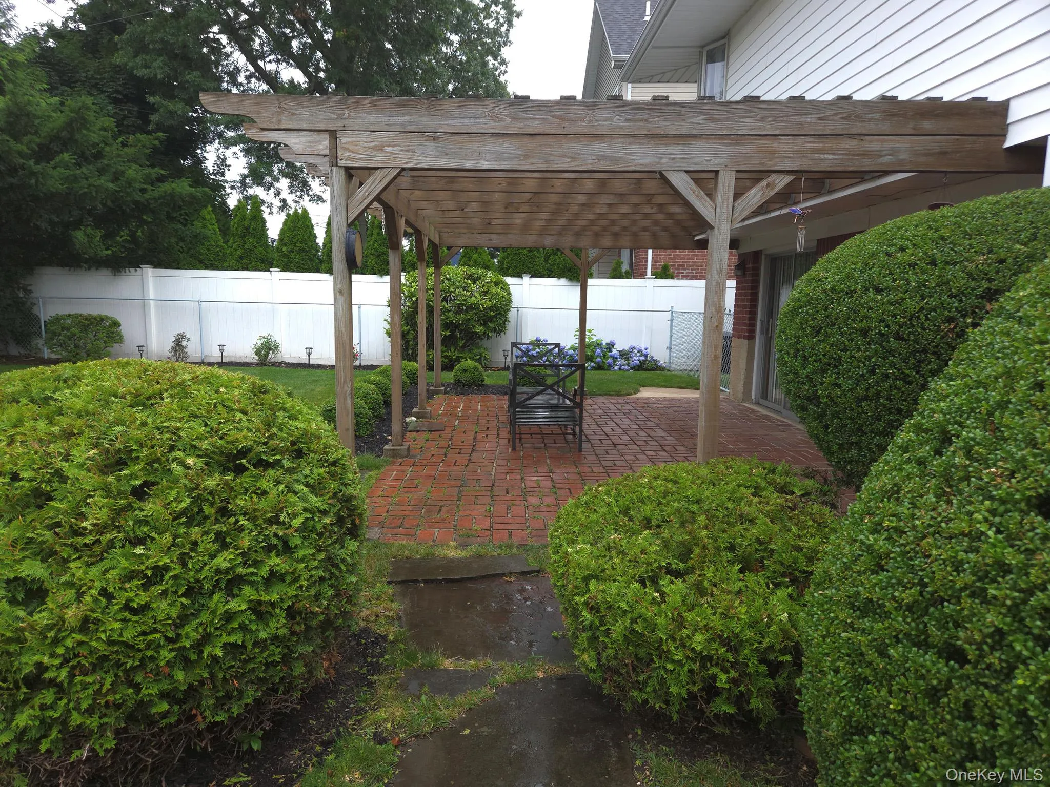 Brick paver patio featuring a wooden pergola with translucent roofing panels, surrounded by a manicured lawn, mature shrubs, and fencing Brick paver patio featuring a wooden pergola with translucent roofing panels, surrounded by a manicured lawn, mature shrubs, and fencing
