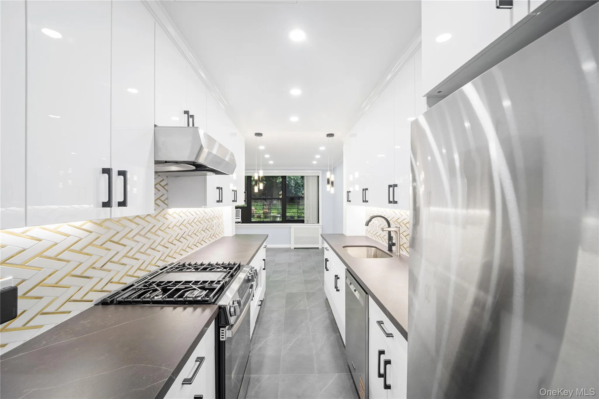 Galley kitchen featuring high-gloss white cabinetry, dark countertops, a herringbone tile backsplash with metallic accents, and stainless steel appliances including a gas range and refrigerator Galley kitchen featuring high-gloss white cabinetry, dark countertops, a herringbone tile backsplash with metallic accents, and stainless steel appliances including a gas range and refrigerator