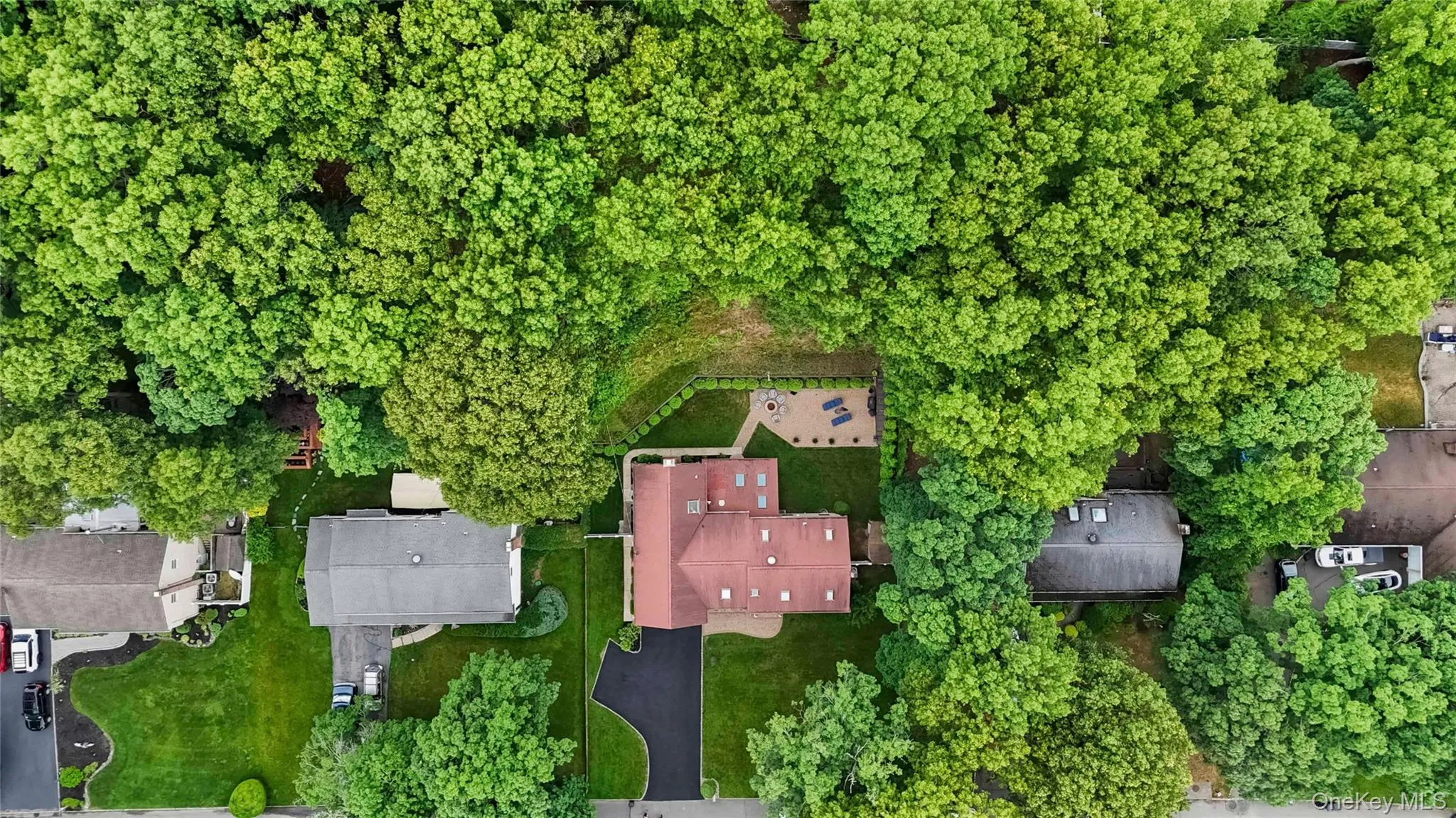 Property surrounded by mature trees, featuring a red-toned roof, multiple skylights, and an expansive paved driveway Property surrounded by mature trees, featuring a red-toned roof, multiple skylights, and an expansive paved driveway