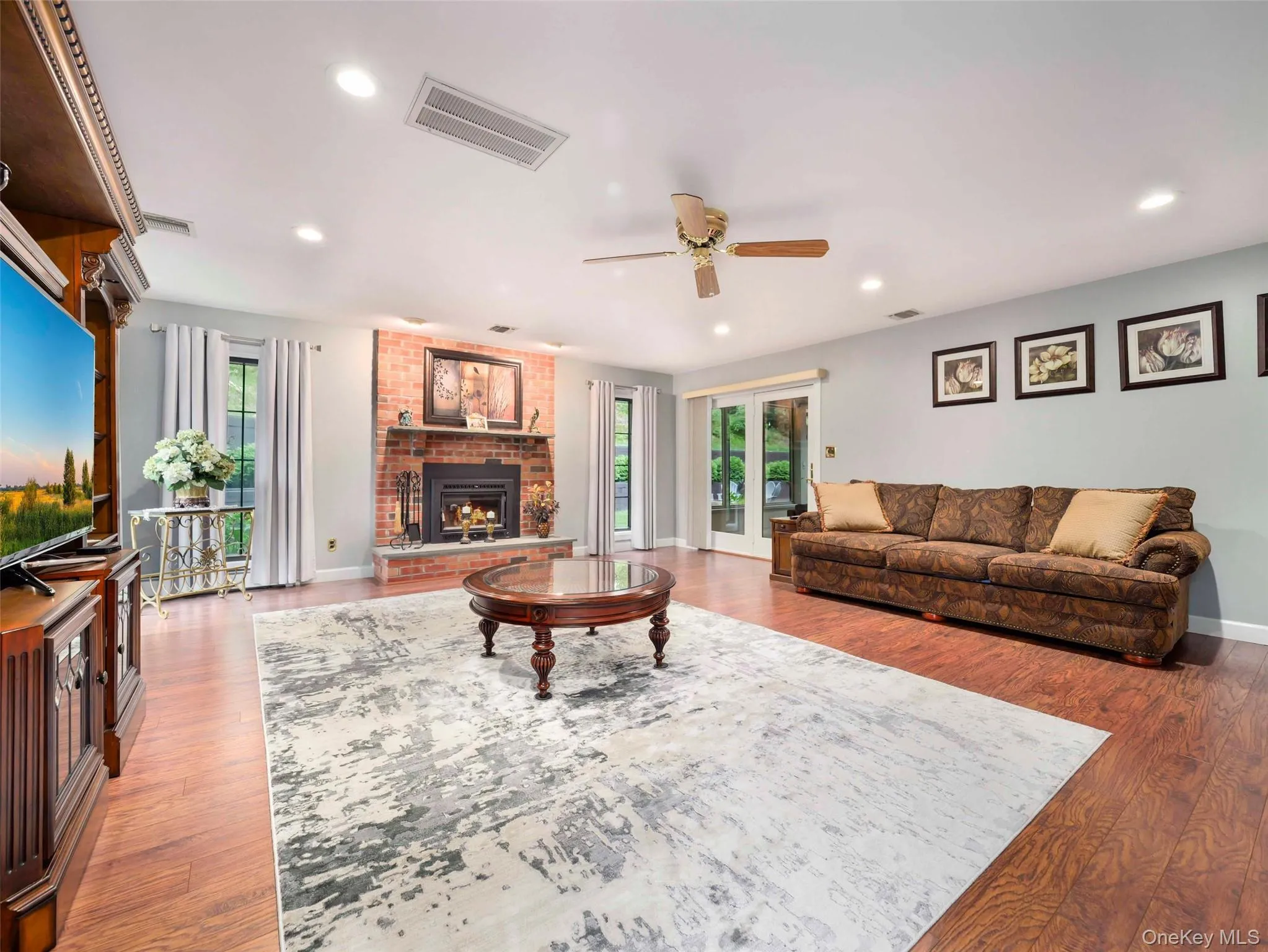 Spacious living area featuring a brick-surround fireplace with a stone hearth, wood-finish flooring, recessed lighting, and a ceiling fan Spacious living area featuring a brick-surround fireplace with a stone hearth, wood-finish flooring, recessed lighting, and a ceiling fan