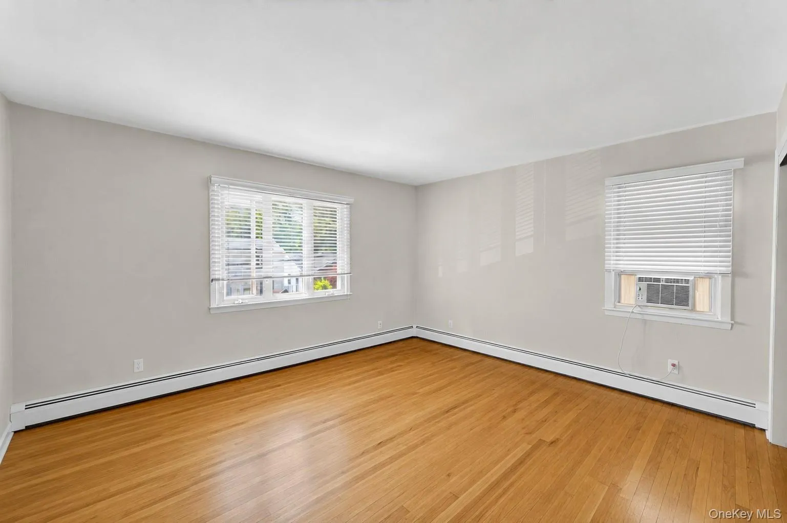 Spacious interior room featuring wood-finish flooring, light-toned walls, two windows with blinds, and baseboard heating Spacious interior room featuring wood-finish flooring, light-toned walls, two windows with blinds, and baseboard heating