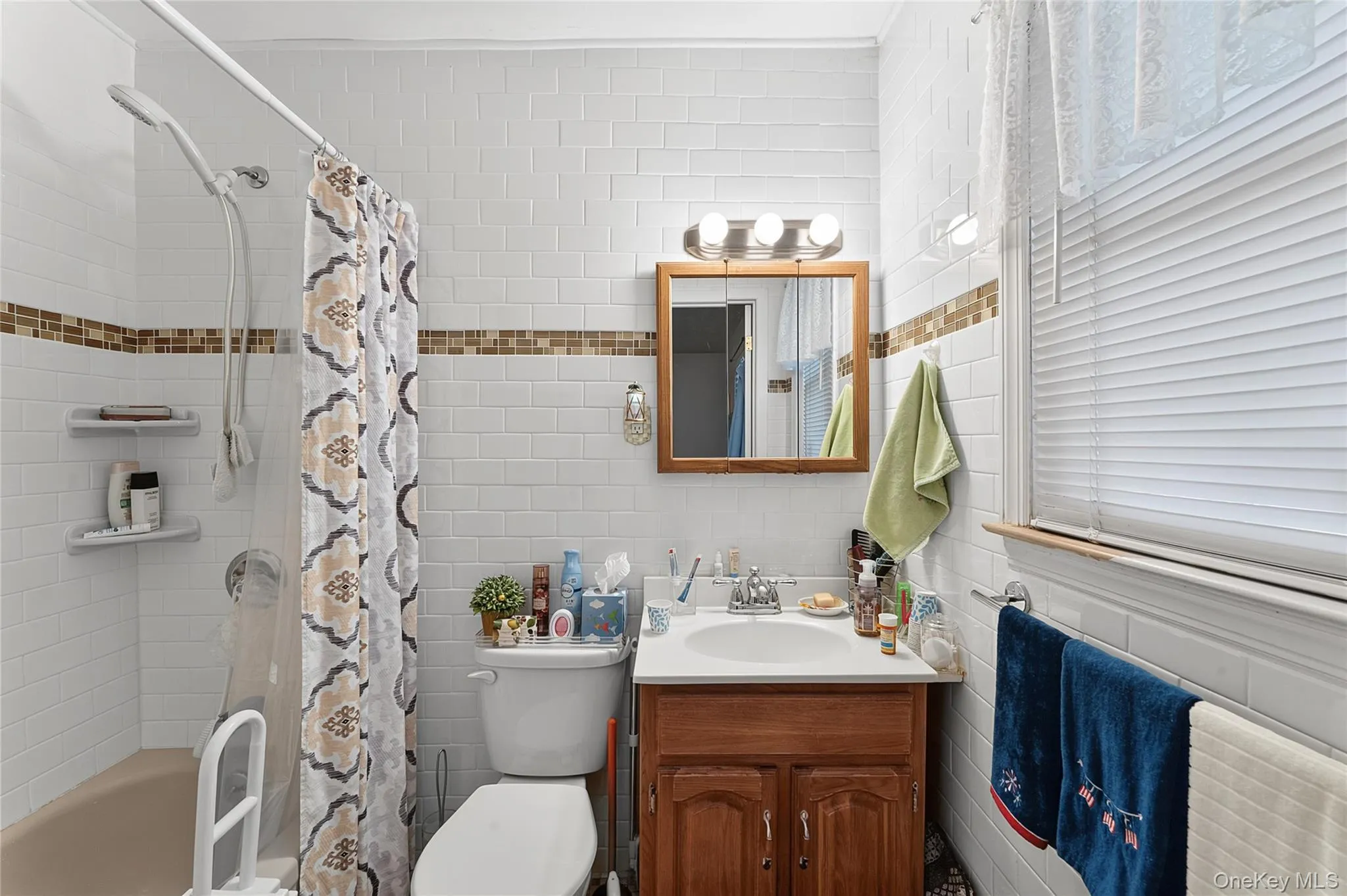 Bathroom featuring white subway tile walls with a decorative mosaic accent band, a built-in bathtub with shower, a single vanity with wood-finish cabinetry, and a chrome light fixture Bathroom featuring white subway tile walls with a decorative mosaic accent band, a built-in bathtub with shower, a single vanity with wood-finish cabinetry, and a chrome light fixture