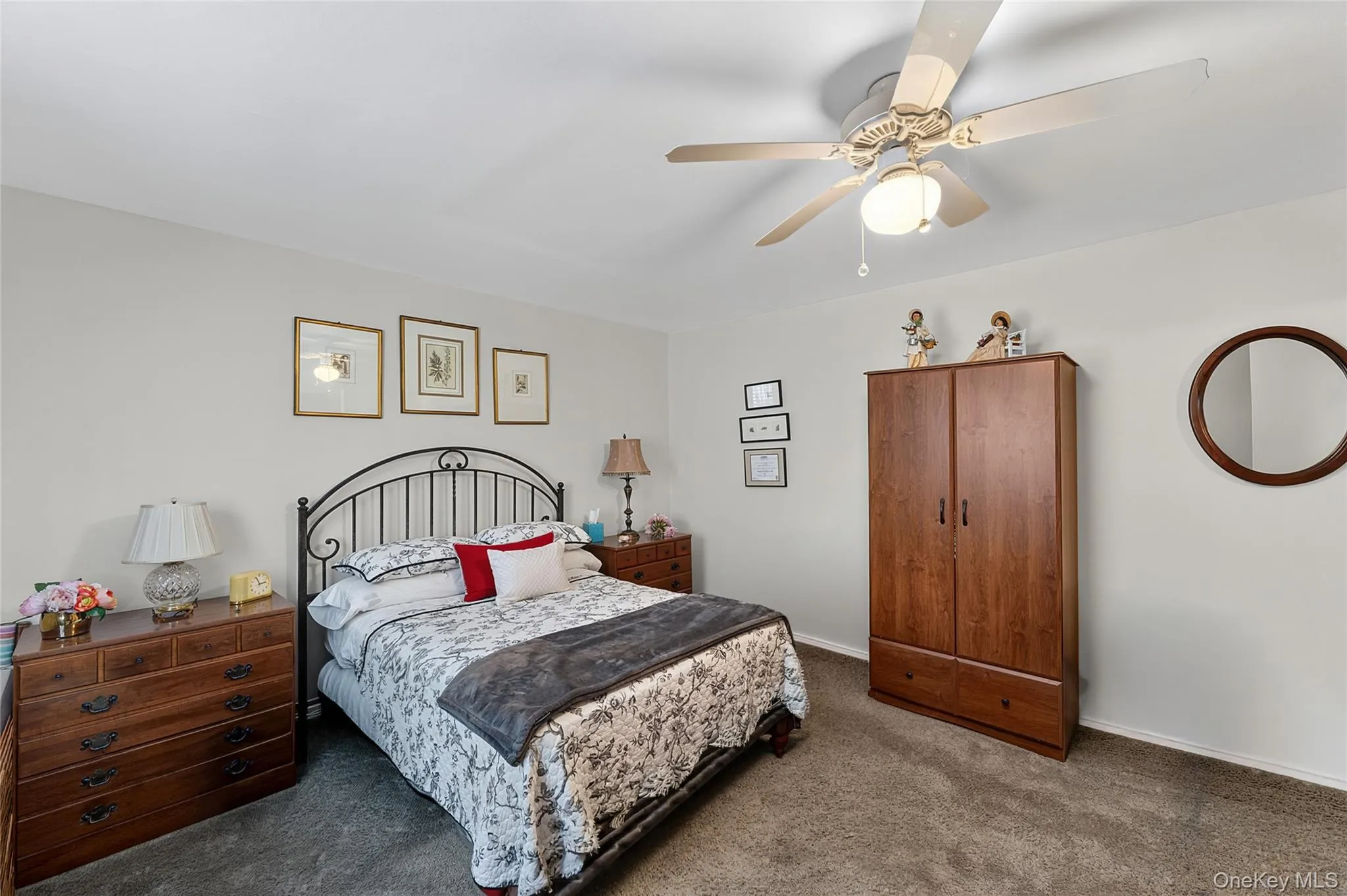 Carpeted bedroom featuring a ceiling fan with integrated lighting, a dark wood-finish armoire, and a rounded wall mirror Carpeted bedroom featuring a ceiling fan with integrated lighting, a dark wood-finish armoire, and a rounded wall mirror