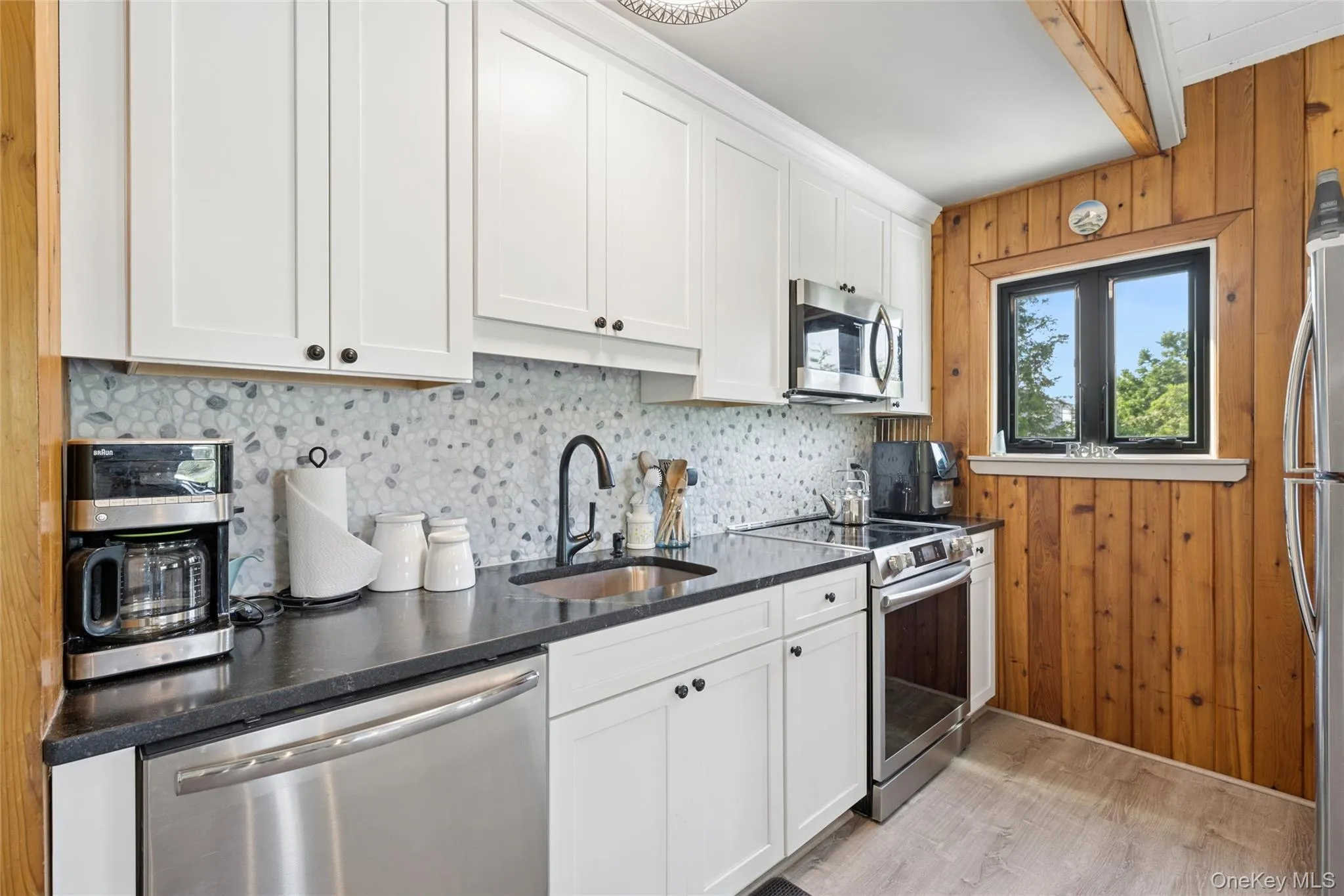Modern kitchen featuring white shaker cabinetry, stainless steel appliances, dark countertops, a pebble-style backsplash, and wood-finish flooring Modern kitchen featuring white shaker cabinetry, stainless steel appliances, dark countertops, a pebble-style backsplash, and wood-finish flooring