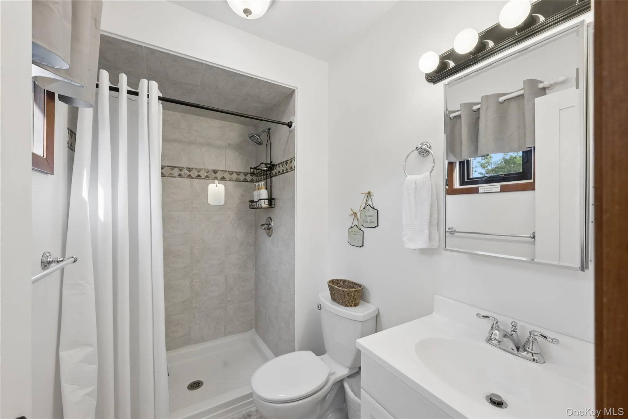 Bathroom featuring a stand-up shower with tiled walls and decorative accent band, white vanity with integrated sink, chrome faucet, wall-mounted mirror, and overhead vanity lighting Bathroom featuring a stand-up shower with tiled walls and decorative accent band, white vanity with integrated sink, chrome faucet, wall-mounted mirror, and overhead vanity lighting