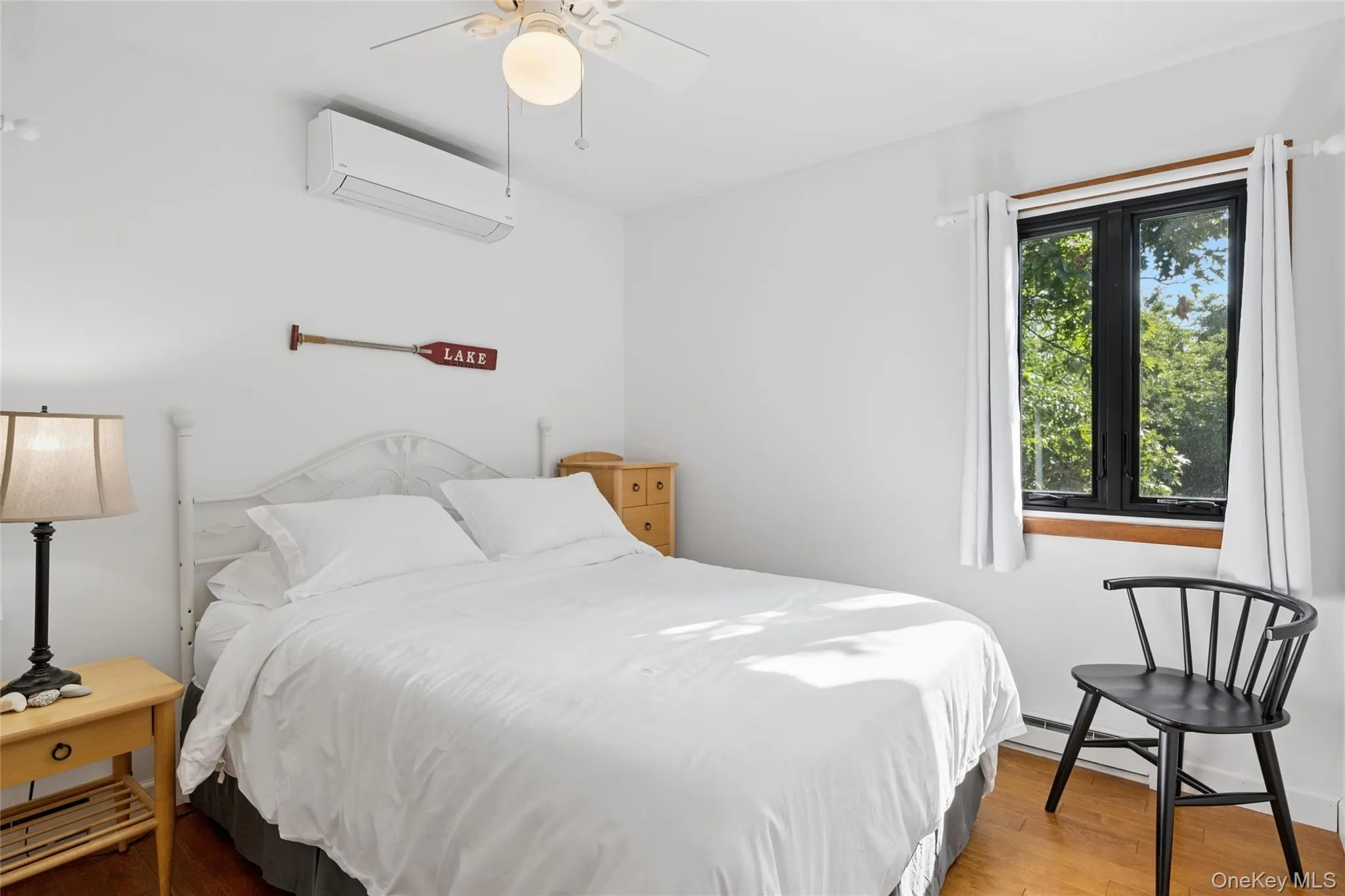 Bedroom featuring a ceiling fan, a wall-mounted air conditioning unit, a large window with black framing, and wood-finish flooring Bedroom featuring a ceiling fan, a wall-mounted air conditioning unit, a large window with black framing, and wood-finish flooring