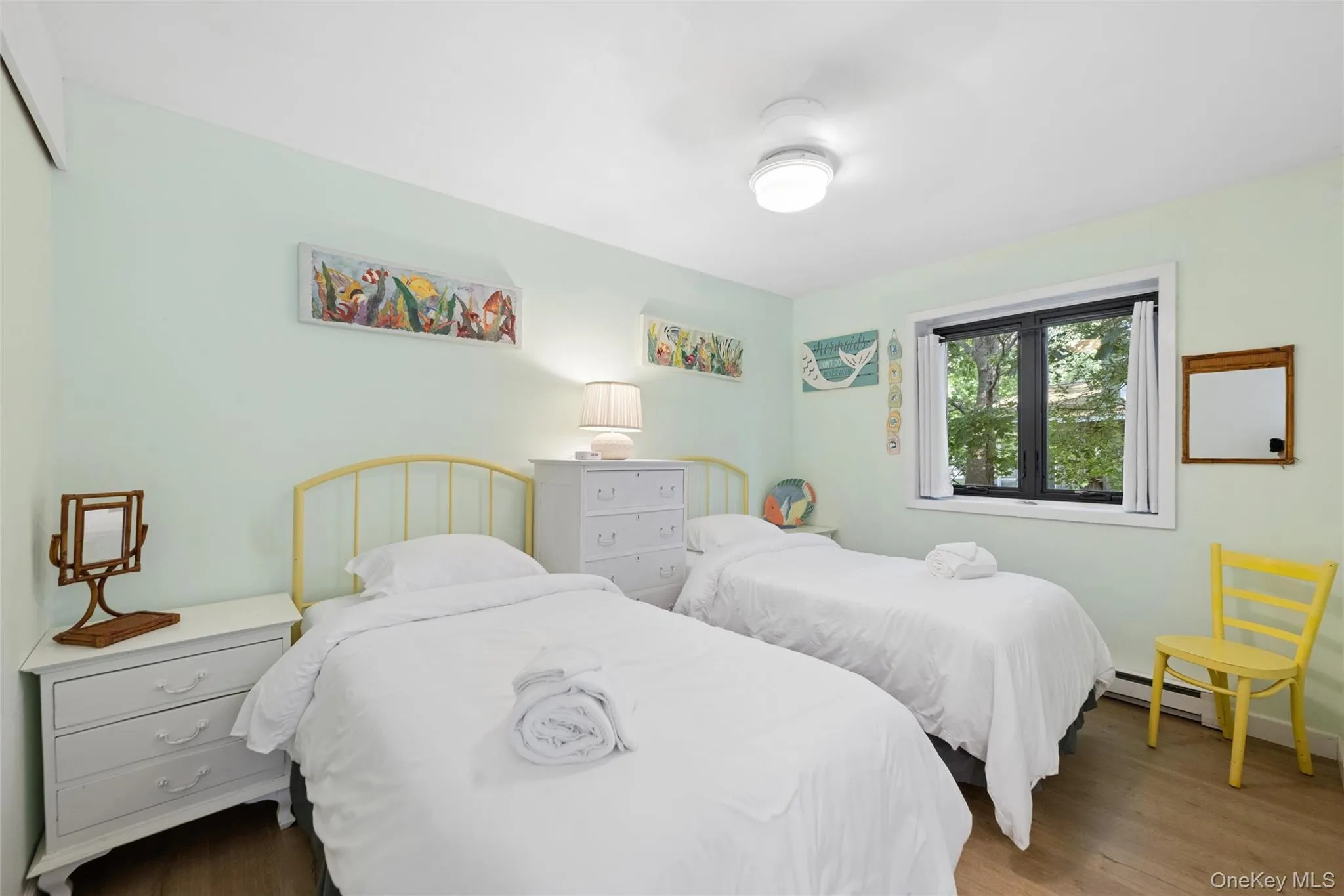 Interior room featuring light green walls, wood-finish flooring, a window with dark trim, white baseboards, and a ceiling-mounted light fixture Interior room featuring light green walls, wood-finish flooring, a window with dark trim, white baseboards, and a ceiling-mounted light fixture