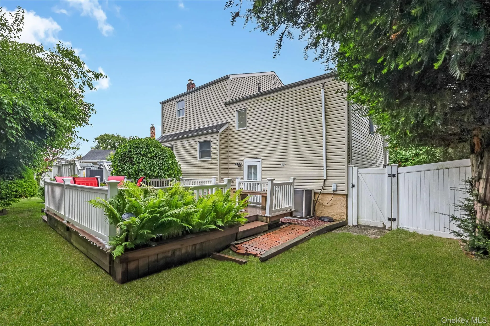 Expansive backyard featuring a raised wooden deck with white railings, integrated planter boxes with greenery, a brick paver pathway, and a privacy fence Expansive backyard featuring a raised wooden deck with white railings, integrated planter boxes with greenery, a brick paver pathway, and a privacy fence