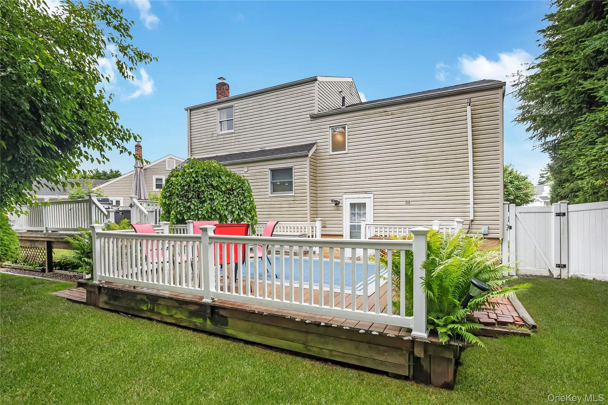 Expansive backyard featuring a raised wooden deck with white railings, integrated planter boxes with greenery, a brick paver pathway, and a privacy fence Expansive backyard featuring a raised wooden deck with white railings, integrated planter boxes with greenery, a brick paver pathway, and a privacy fence