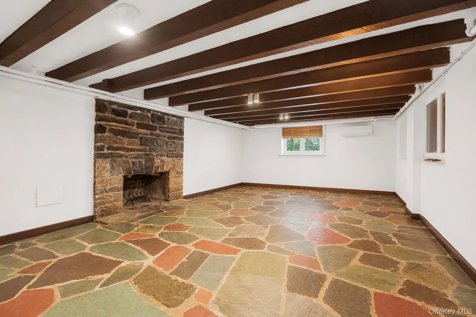 Garden-level recreation room with a stone fireplace, exposed beam ceiling, and flagstone flooring offering exceptional space for entertaining or recreation.Virtually enhanced to remove personal items. Garden-level recreation room with a stone fireplace, exposed beam ceiling, and flagstone flooring offering exceptional space for entertaining or recreation.Virtually enhanced to remove personal items.