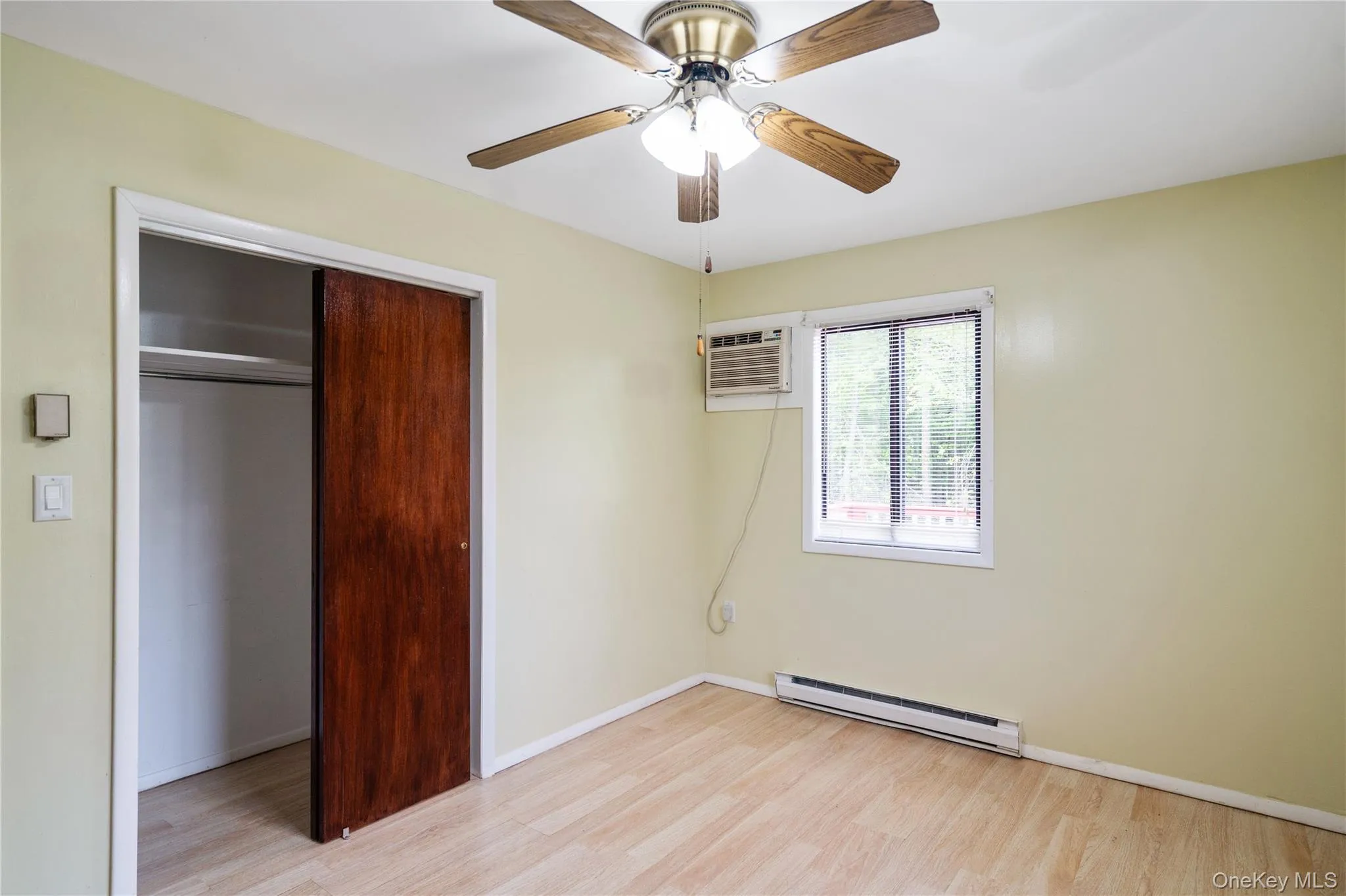 Unfurnished bedroom with a baseboard radiator, light wood-type flooring, a closet, a wall mounted AC, and baseboards Unfurnished bedroom with a baseboard radiator, light wood-type flooring, a closet, a wall mounted AC, and baseboards