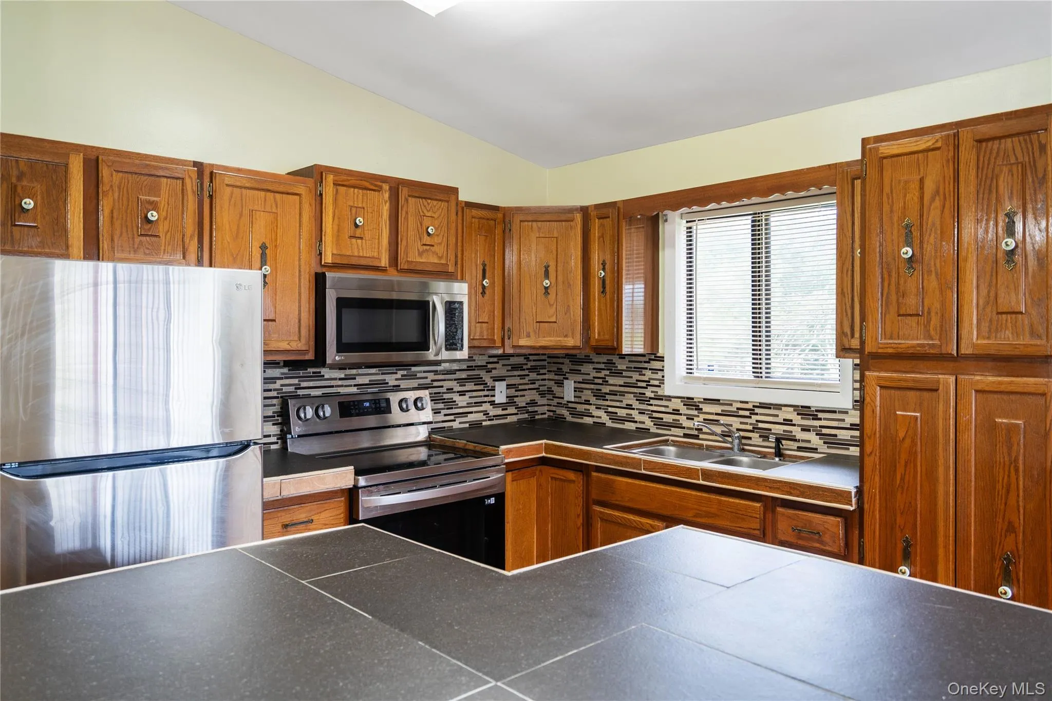 Kitchen with appliances with stainless steel finishes, lofted ceiling, tasteful backsplash, brown cabinets, and dark countertops Kitchen with appliances with stainless steel finishes, lofted ceiling, tasteful backsplash, brown cabinets, and dark countertops