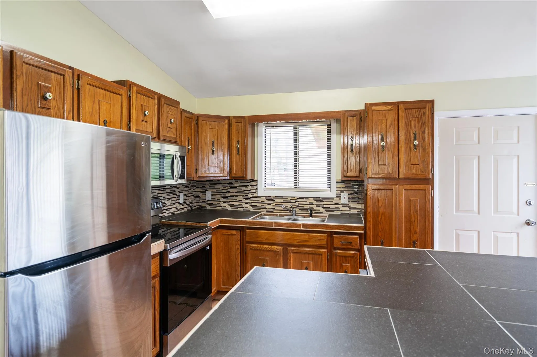 Kitchen featuring appliances with stainless steel finishes, a sink, brown cabinetry, tasteful backsplash, and vaulted ceiling Kitchen featuring appliances with stainless steel finishes, a sink, brown cabinetry, tasteful backsplash, and vaulted ceiling