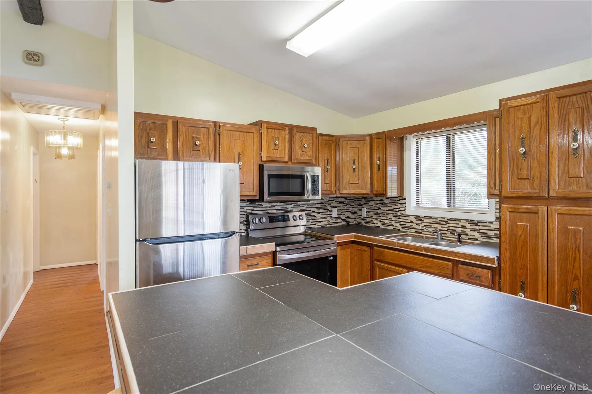 Kitchen with appliances with stainless steel finishes, lofted ceiling, dark countertops, tasteful backsplash, and brown cabinetry Kitchen with appliances with stainless steel finishes, lofted ceiling, dark countertops, tasteful backsplash, and brown cabinetry