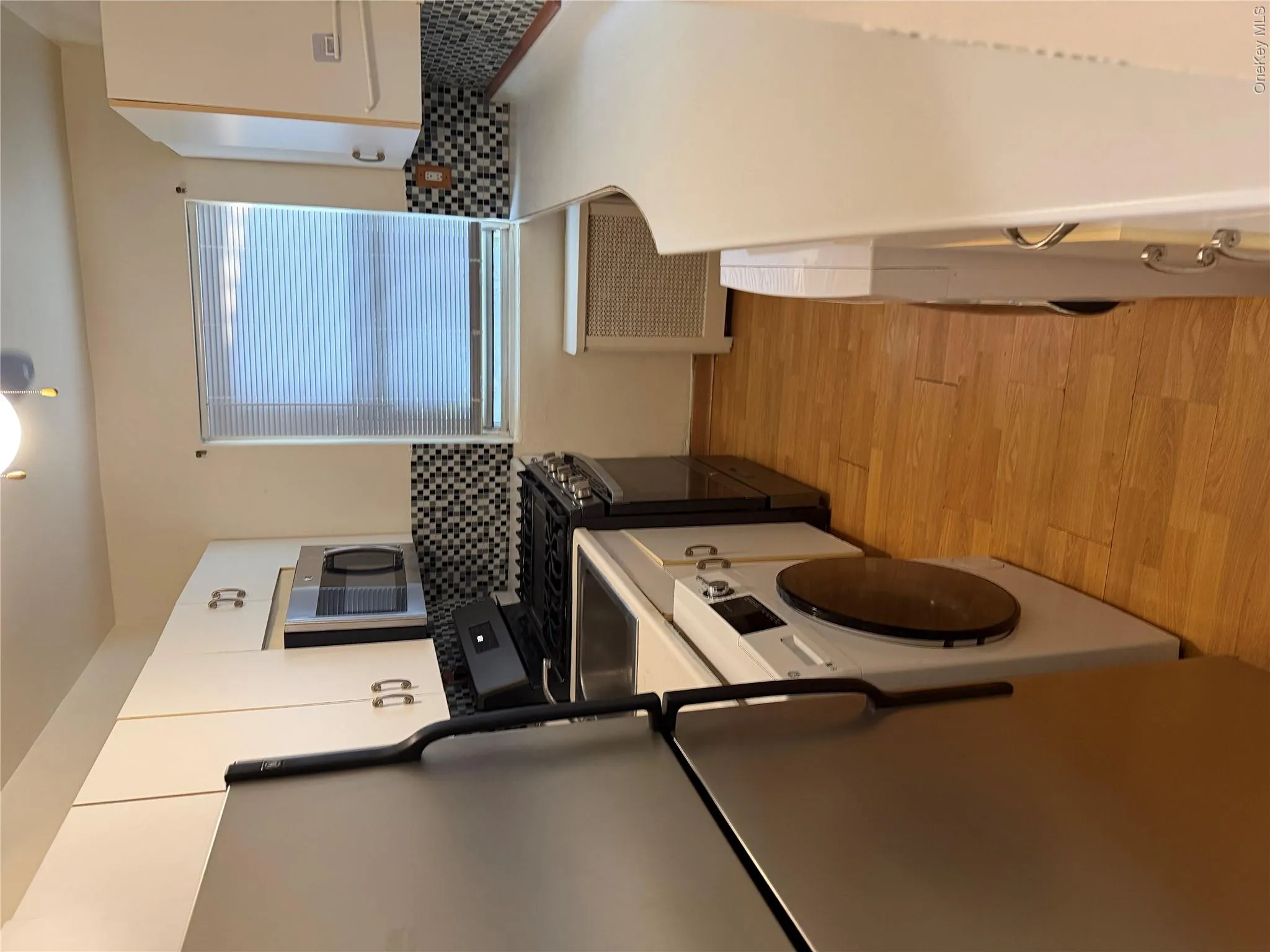 Galley kitchen featuring wood-finish flooring, white cabinetry with silver-tone hardware, stainless steel appliances, and a mosaic tile backsplash Galley kitchen featuring wood-finish flooring, white cabinetry with silver-tone hardware, stainless steel appliances, and a mosaic tile backsplash