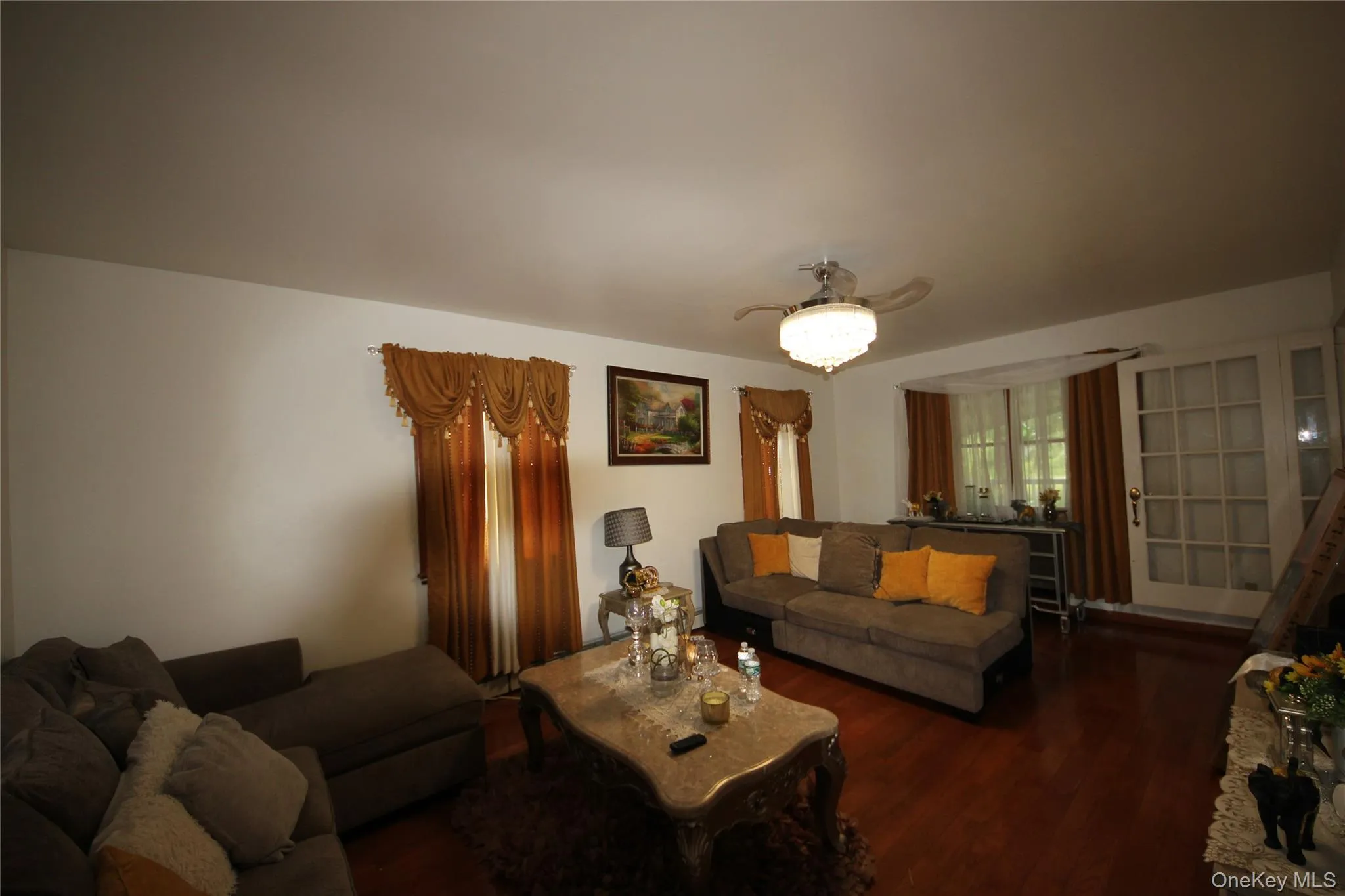 Spacious living area featuring wood-finish flooring, white walls, and a ceiling fan with integrated lighting Spacious living area featuring wood-finish flooring, white walls, and a ceiling fan with integrated lighting