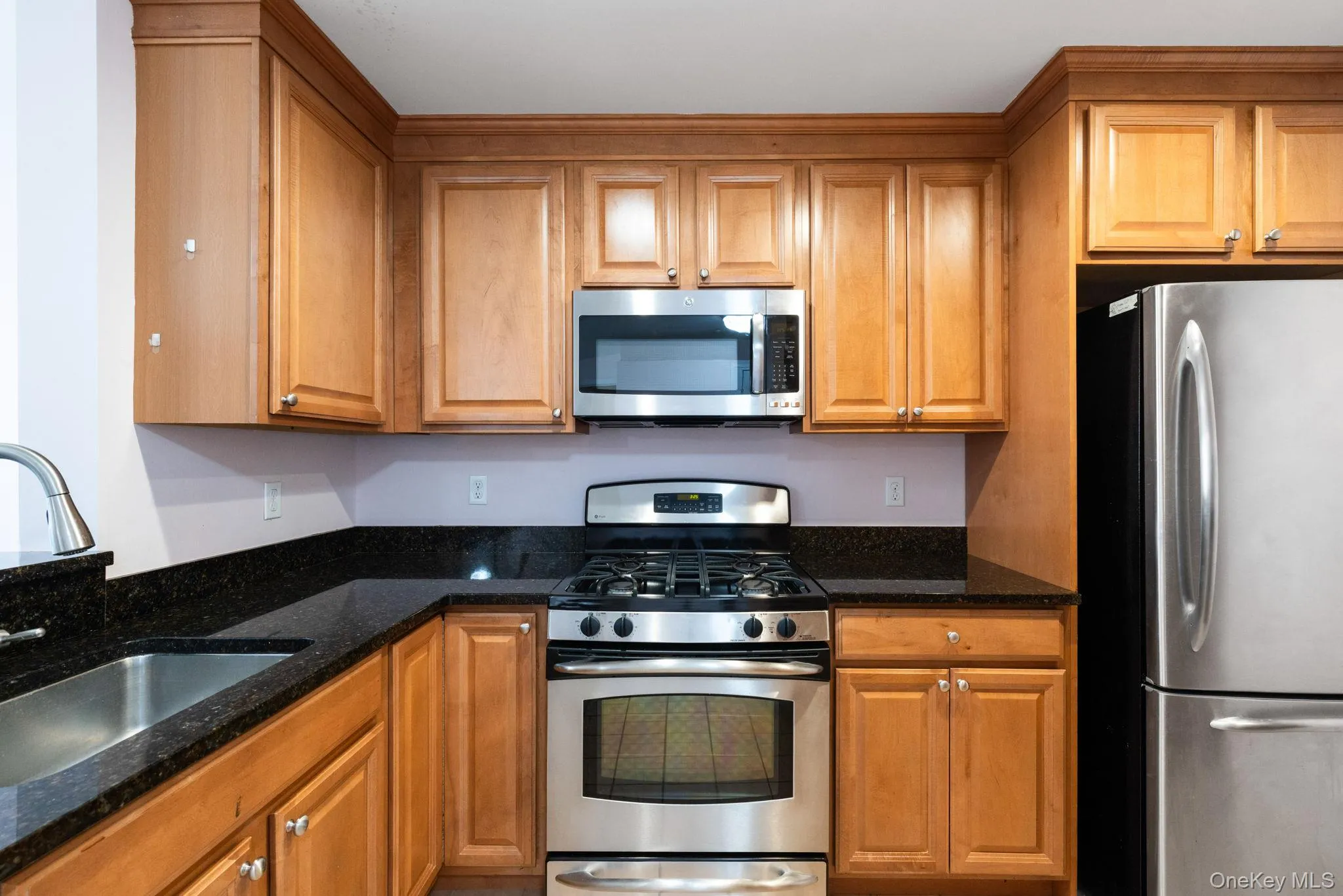 Kitchen with wood-finish cabinetry, dark countertops, a stainless steel gas range, a stainless steel microwave, and a stainless steel refrigerator Kitchen with wood-finish cabinetry, dark countertops, a stainless steel gas range, a stainless steel microwave, and a stainless steel refrigerator