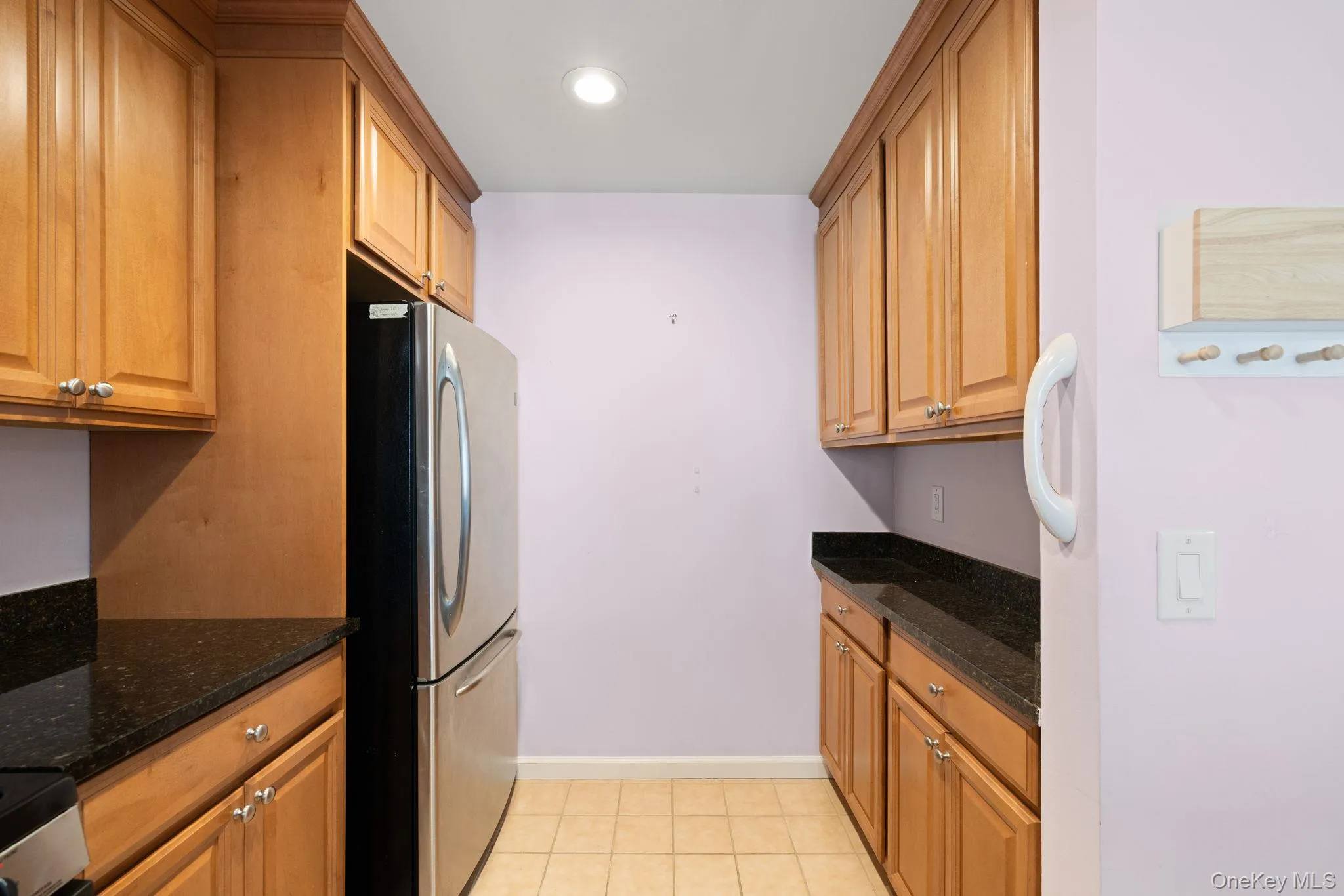 Galley kitchen featuring wood-finish cabinetry, dark stone countertops, stainless steel refrigerator, recessed lighting, and tile flooring Galley kitchen featuring wood-finish cabinetry, dark stone countertops, stainless steel refrigerator, recessed lighting, and tile flooring