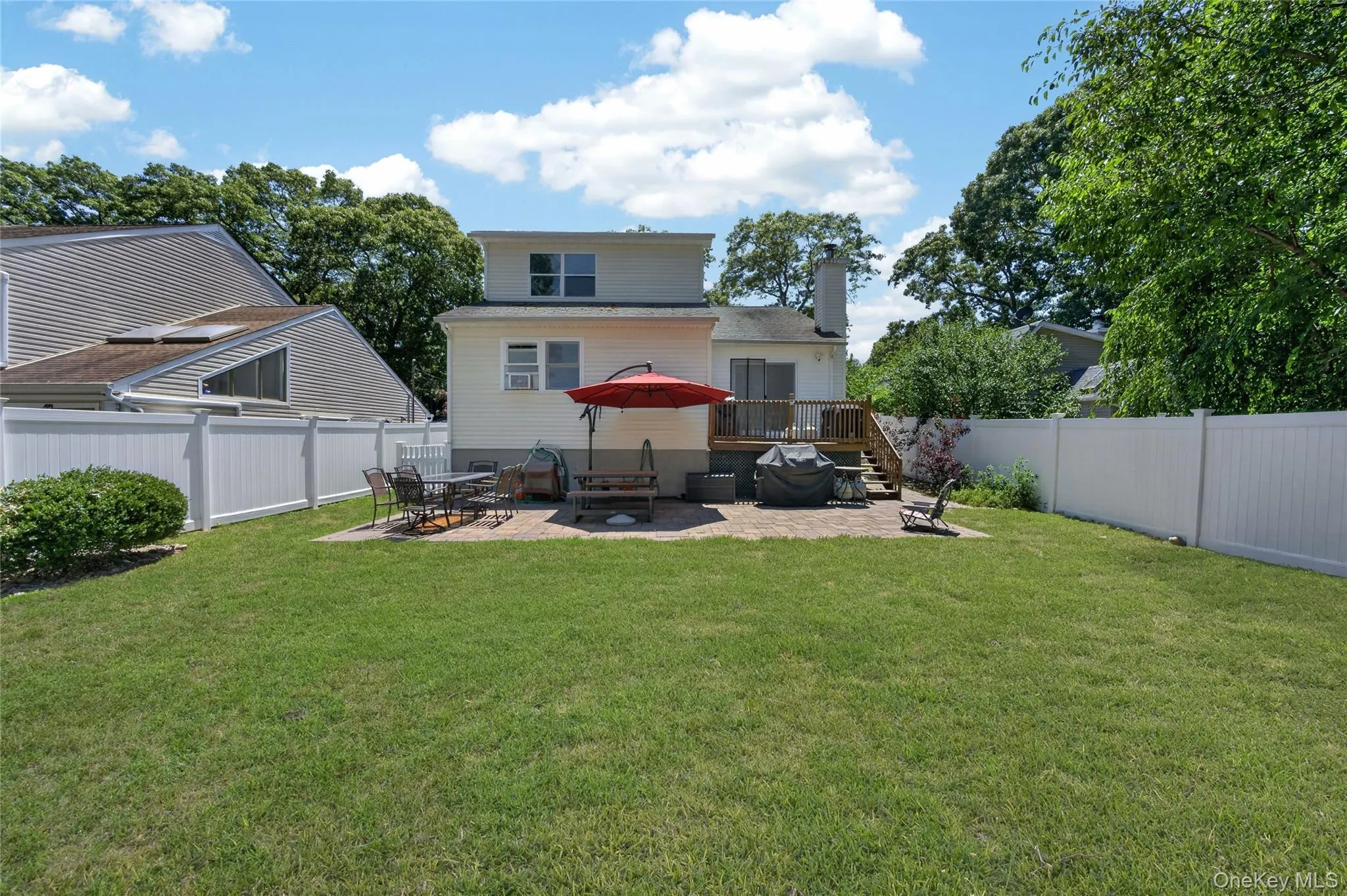 Spacious backyard featuring a manicured lawn, paver patio, wood deck, and white vinyl perimeter fencing Spacious backyard featuring a manicured lawn, paver patio, wood deck, and white vinyl perimeter fencing